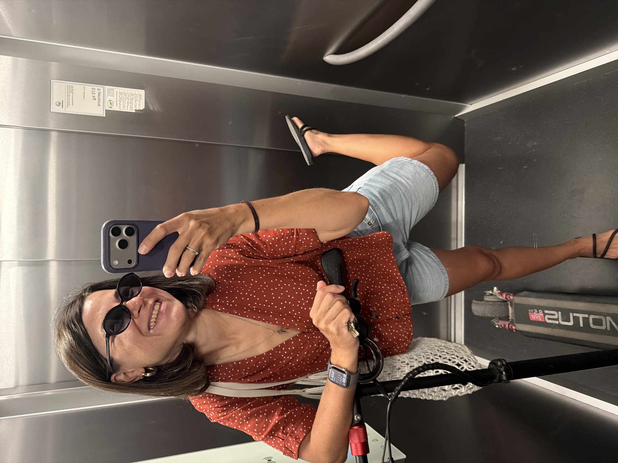The image shows a selfie of a smiling woman taken inside an elevator in Budva, Montenegro. The photograph is rotated 90 degrees clockwise, so the woman appears on her side. She has medium-length brown hair and is wearing sunglasses, a red short-sleeved top with white polka dots, light blue denim shorts, and black flip-flops. She holds a smartphone with a purple case in her left hand to capture the image, while her right hand rests on the handlebar of a black electric scooter positioned beside her. A white net bag hangs from the scooter's handle. The woman also wears a watch on her left wrist and a ring on her left ring finger. The elevator features metallic walls and a grab bar. Visible text includes a mirrored "a belGouR" on a sticker affixed to the elevator wall, "2UTON," "2.8," and "019" on the side of the scooter, and numbers such as "8:48" and "60" on her smartwatch display.