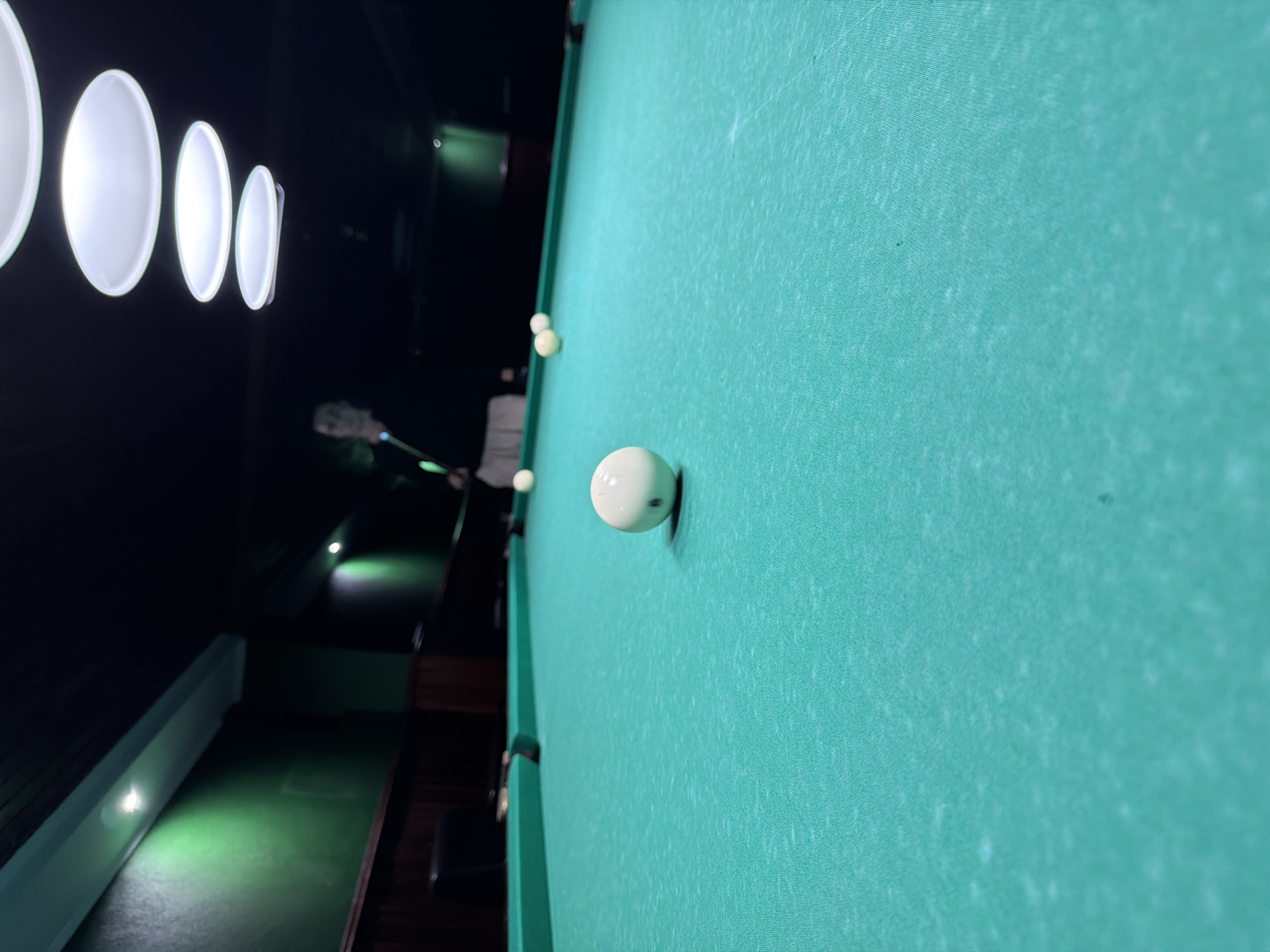 The image shows a close-up view of a pool table covered in bright green felt. A white cue ball is positioned in the foreground, slightly off-center to the right. Several other billiard balls are scattered across the table in the mid-ground and background, some of which are white. In the background, a person is partially visible, holding a pool cue and appearing to be playing the game. The environment is dimly lit, with overhead lights casting a soft glow on the table. The walls of the room are dark green, and wooden paneling is visible along the lower sections. The overall impression is of an indoor billiard hall or game room. There is no specific information available about the location in the city of Strășeni, Moldova.