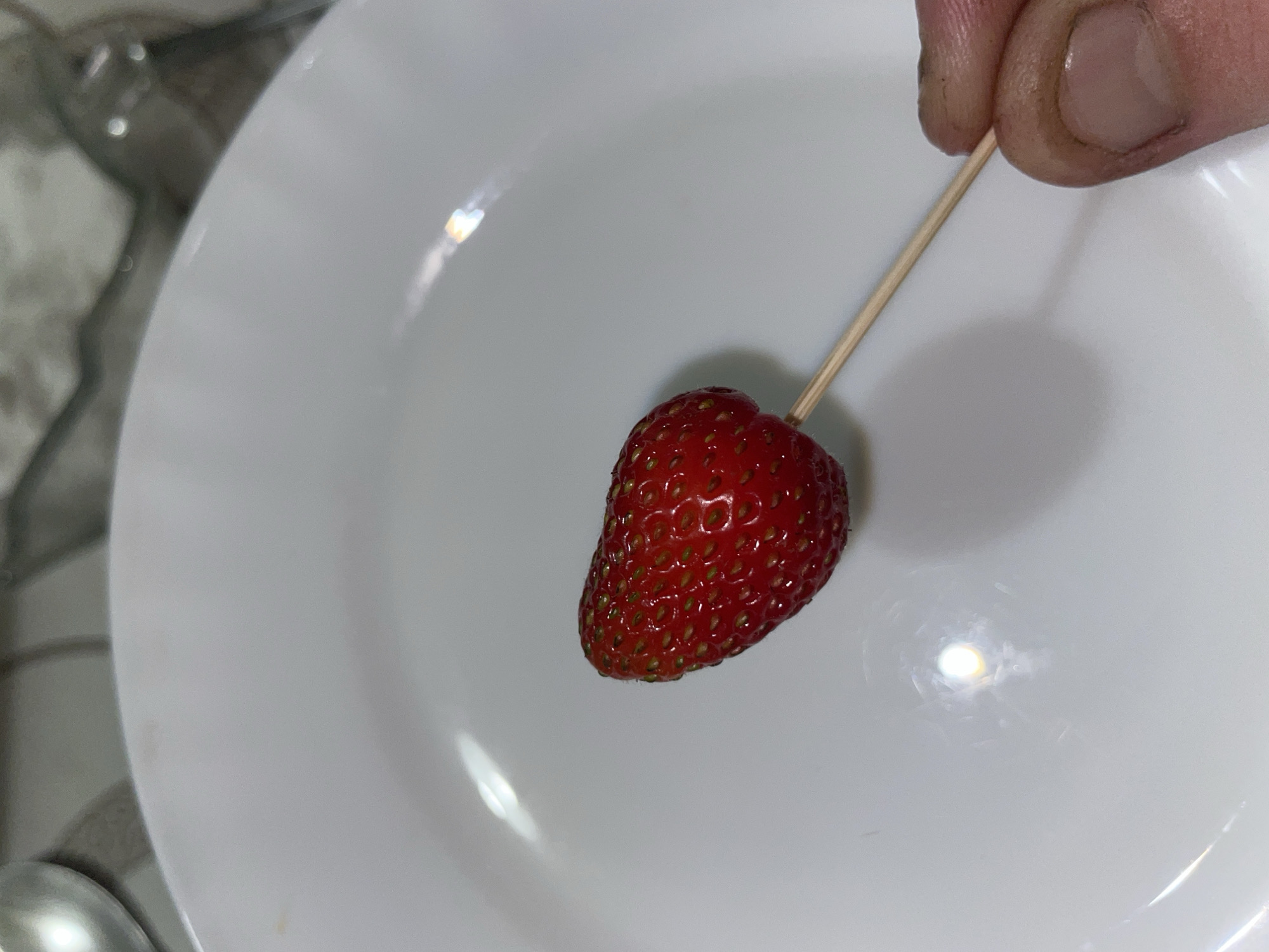 A single strawberry, impaled on a wooden skewer, rests on a white plate. The strawberry is red and plump, with visible seeds. A person's hand, with dirty fingernails, is holding the skewer. The background is blurred but suggests an indoor setting with a countertop and possibly some kitchen utensils. The image was taken in Laishevo, Russia.