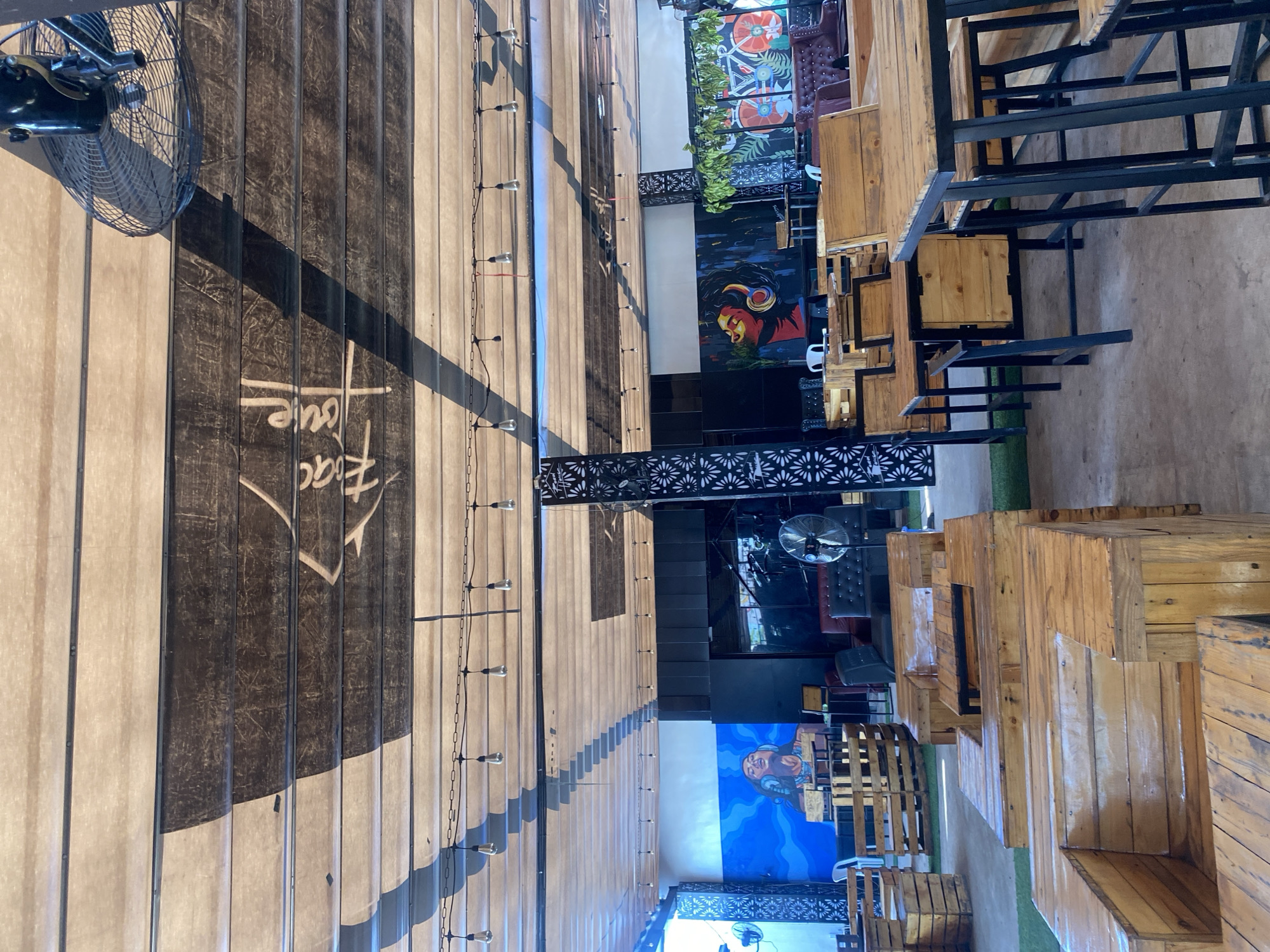 This image captures the interior of an unoccupied open-air or semi-enclosed establishment, likely a cafe or lounge, located in Howayi, Nigeria. The overhead structure is comprised of alternating light beige and dark brown horizontal panels, consistent with a retractable awning system, on which the inverted white text "Frac House" is visible. Multiple strands of string lights with exposed Edison-style bulbs are suspended across the ceiling. A black industrial-style ceiling fan is prominently positioned in the upper right. The space is furnished with various items made from light-toned wood, notably repurposed pallets, forming low seating arrangements (benches and armchairs) and tables in the foreground and mid-ground. Taller wooden tables with black metal supports and matching stools are also present. The floor consists of concrete interspersed with sections of artificial green turf. The back walls feature large-scale murals; one on the left depicts a figure with headphones on a blue background, and another on the right showcases a similar subject on an abstract dark background. Further right, a mural featuring a bicycle and floral patterns is visible. Black decorative screens with a repetitive floral motif serve as architectural accents or dividers. Additional dark upholstered seating and freestanding fans are discernible in the background. No individuals are present in the scene.