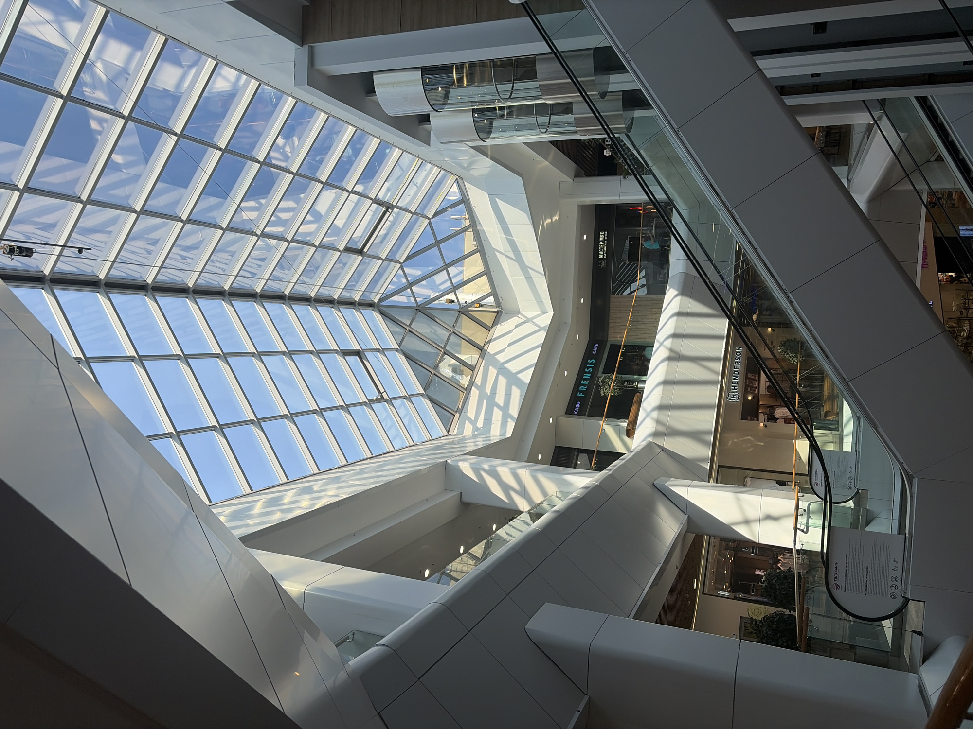 The image depicts the interior of a multi-level shopping mall. A large, geometric glass ceiling composed of numerous rectangular panes illuminates the space with natural light. The sky is visible through the glass panes. White structural beams and supports are a prominent feature of the architecture. Escalators ascend through the levels of the mall. There is a clear view to the other levels.
Signage in Russian for "KAOE FRENSIS" is visible, along with other store signs, indicating the location is in Russia.
Based on the architectural style and the Russian signage, this image is likely taken in the shopping mall "Taligan" in Izhevsk, Russia.