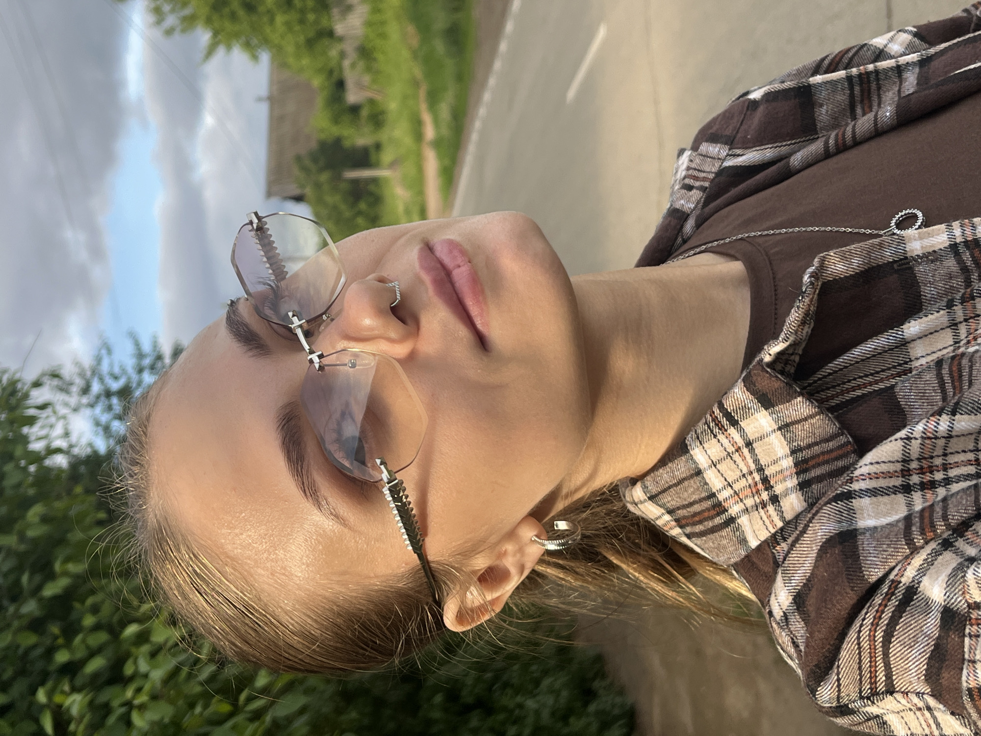 This close-up outdoor portrait features a young person, likely female, positioned with her head tilted back and to the side, gazing upwards and slightly left within the frame. She has light skin, light brown or blonde hair, and wears rimless glasses with distinctive, possibly corrugated, temple arms. Her lips are light pink, and a small silver hoop nose ring is visible. A silver ear cuff adorns her right ear.
She is dressed in a dark brown t-shirt, layered with an unbuttoned plaid shirt featuring shades of brown, beige, and white. A delicate silver chain necklace with a small circular pendant rests on her t-shirt.
The background suggests an outdoor setting in Фоки, Russia. To the lower left of the frame, there is lush green foliage, while the upper right reveals a light-colored road or path and faint outlines of distant structures through trees. The sky is partly cloudy, and the lighting appears soft and warm, characteristic of late afternoon or early evening. The overall atmosphere is relaxed and contemplative. There is no visible text in the image.
