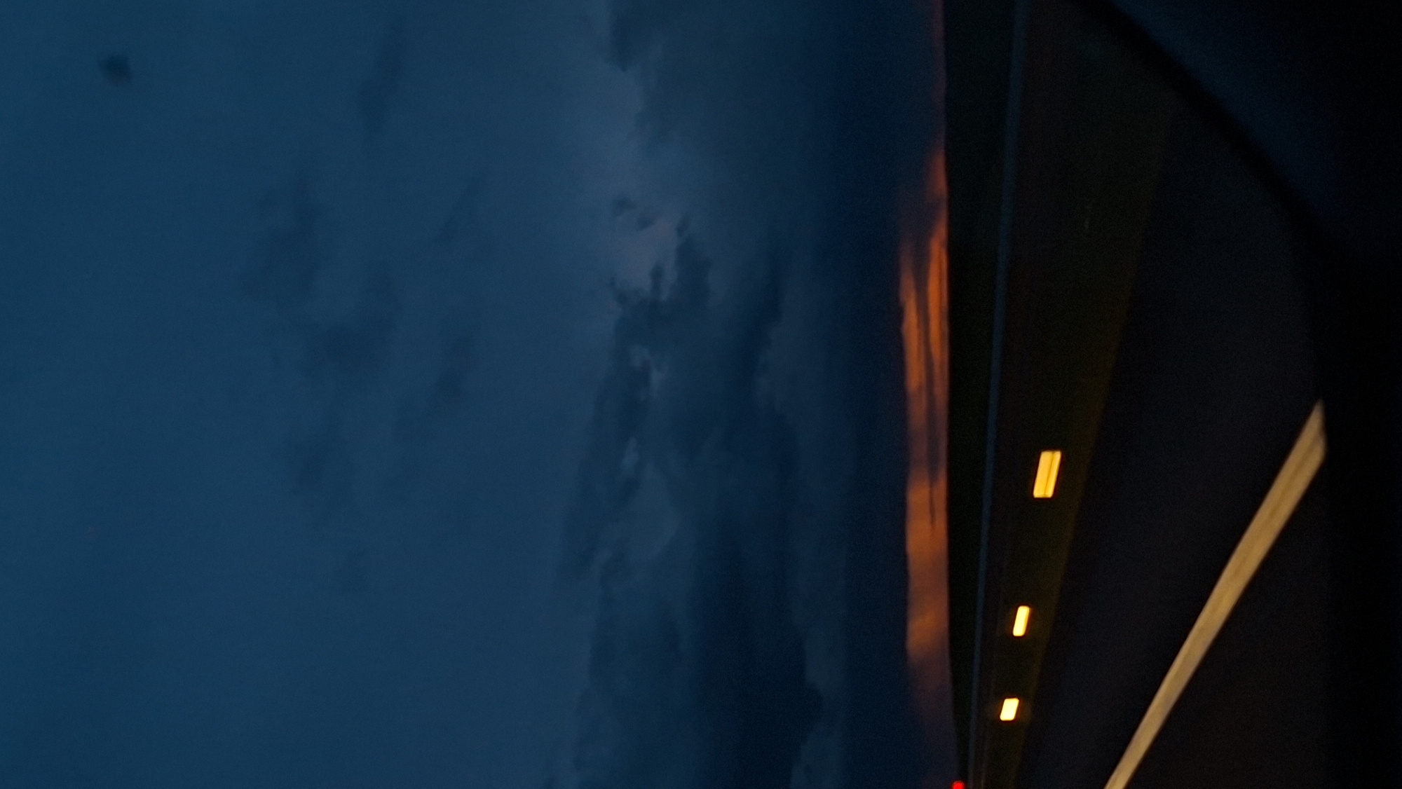 A blurry image, apparently taken from a moving vehicle, shows a portion of a dark, seemingly nighttime sky with cloud formations to the left. A roadside with reflective markers is visible to the right, suggesting a highway or major road. The lighting is low, with the roadside markers reflecting light, indicating artificial illumination. The scene presents an environment likely situated on the outskirts of a city or in a rural area with a connecting highway, but specific details such as people, interaction, or other objects are not discernable given the image's limited scope and blurriness. The exact location within the Дабрынёўскі сельскі Савет is not identifiable.