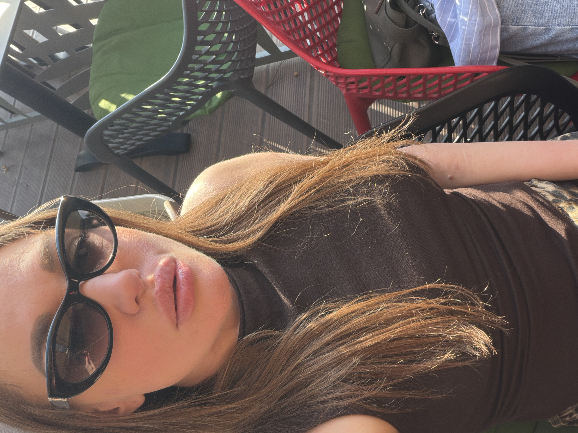The image shows a woman with long brown hair, wearing sunglasses and a brown top. She is sitting outside on a wooden deck, likely at a cafe or restaurant, as indicated by the presence of multiple chairs, some with green cushions and others in red and black. The background includes what appears to be a bag and clothing, suggesting the setting is a social gathering or outdoor dining area. There is no explicit indication this image was taken in Bar, Montenegro, it is assumed based on the query.