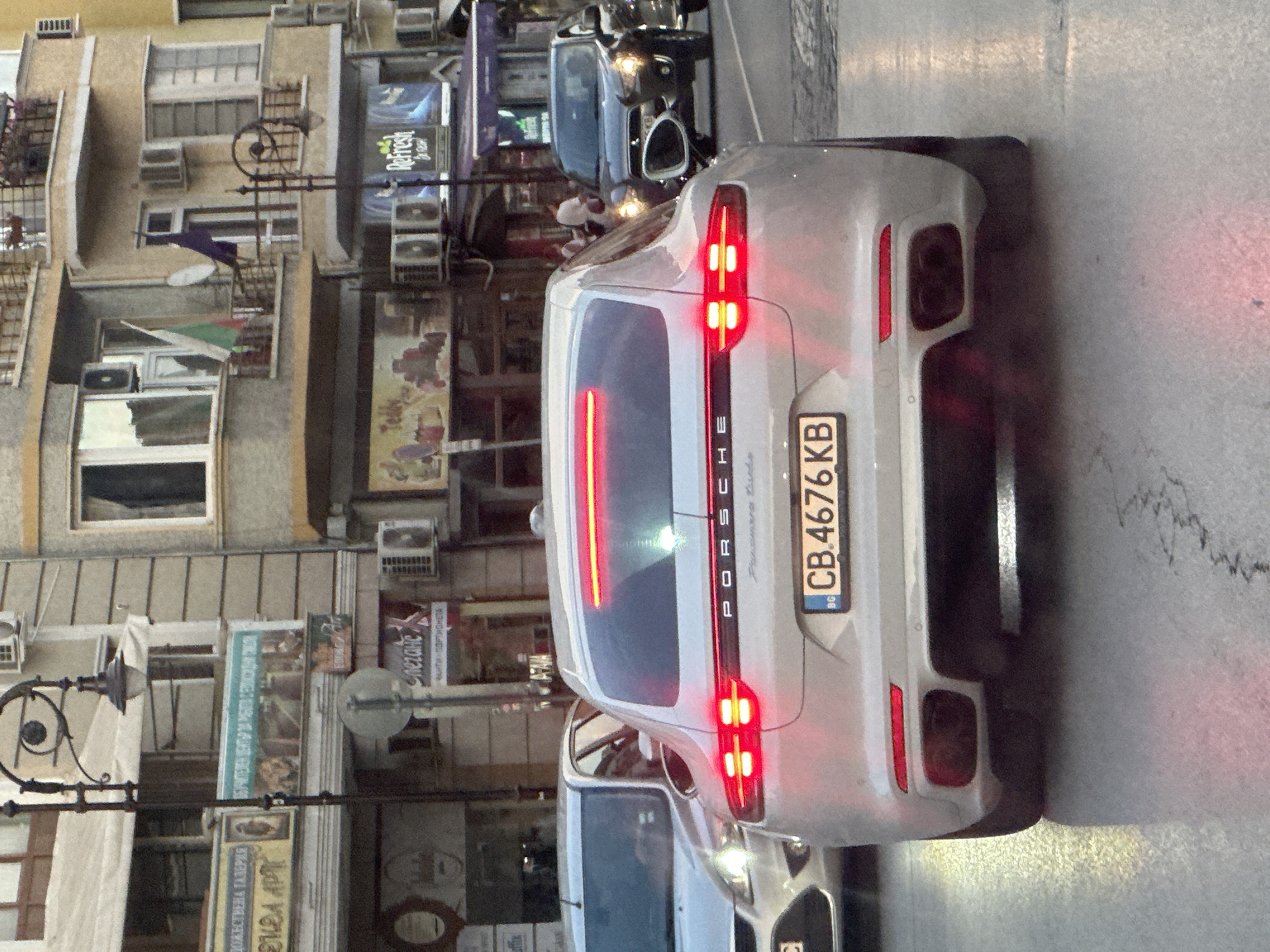 The image captures a street scene in Veliko Tarnovo, Bulgaria, likely during twilight or early evening. The focal point is the rear of a light-colored Porsche Panamera, its taillights illuminated and reflecting on the wet asphalt. The car's license plate reads "CB.4676 KB" and features the Bulgarian flag.
To the left of the car, a multi-story building displays various signs and architectural features. One sign is for "Refresh," and another appears to be a cultural or art gallery, indicated by the text "ПОЖЕСТВЕНА ГАЛЕРИЯ" (POZHESTVENA GALERIYA) and "ΕΚΘΑ ΑΡΓ" (EKTHA ARG), suggesting an art exhibition. The building has numerous windows and air conditioning units. A street lamp with a decorative circular element is also visible.
In the background, other vehicles are present, including a dark-colored car. The lighting suggests it is either dawn or dusk, with artificial lights from the buildings and vehicles contributing to the overall illumination. The wet road surface implies recent rain or damp conditions. The scene appears to be a typical urban environment with shops and residential buildings lining the street. No people are clearly discernible in the image.