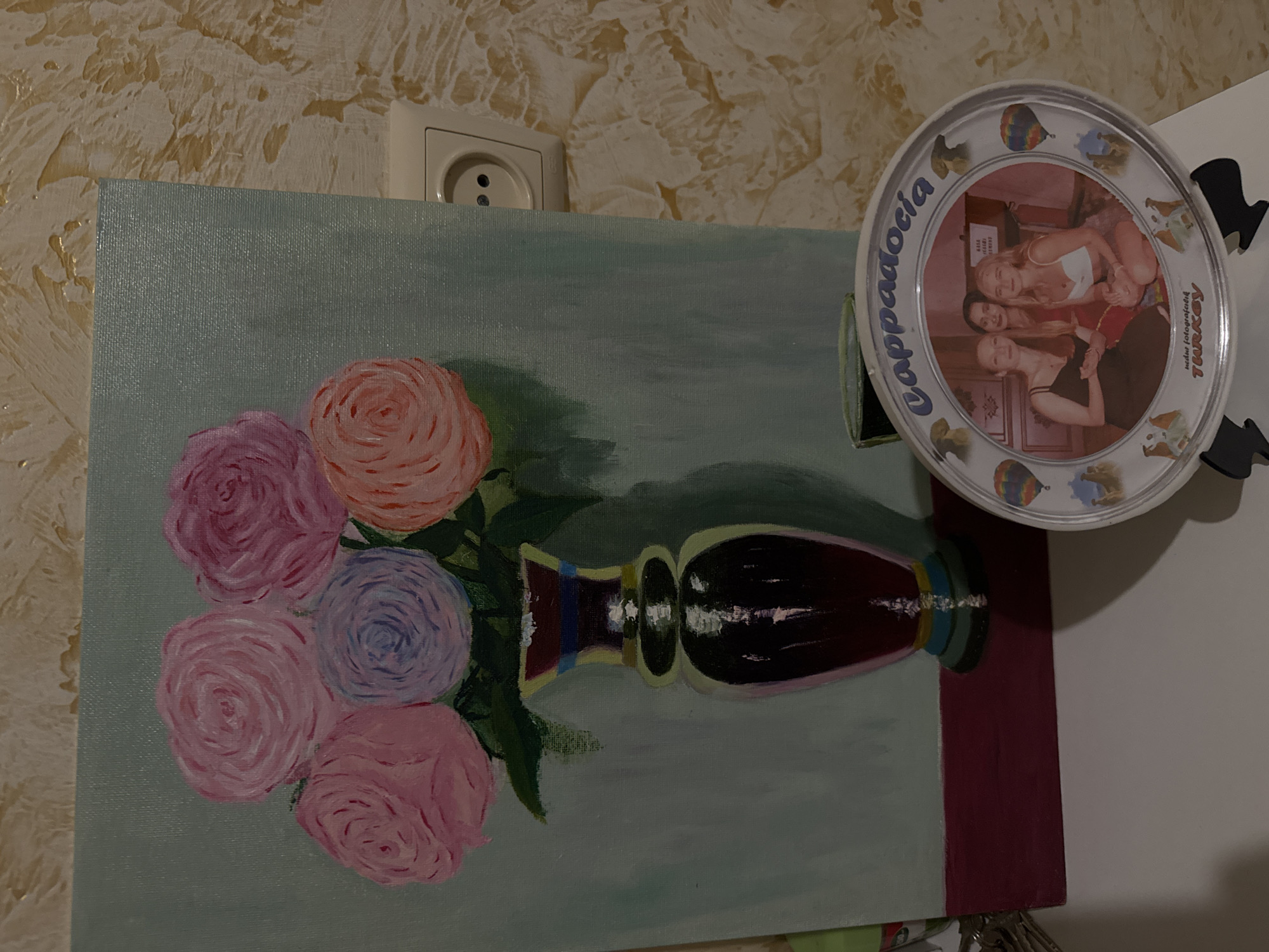 The image shows a painting of flowers in a vase against a wall with a power outlet. A souvenir plate with a photo of three women and the words "Cappadocia, Turkey" is positioned to the right of the painting.