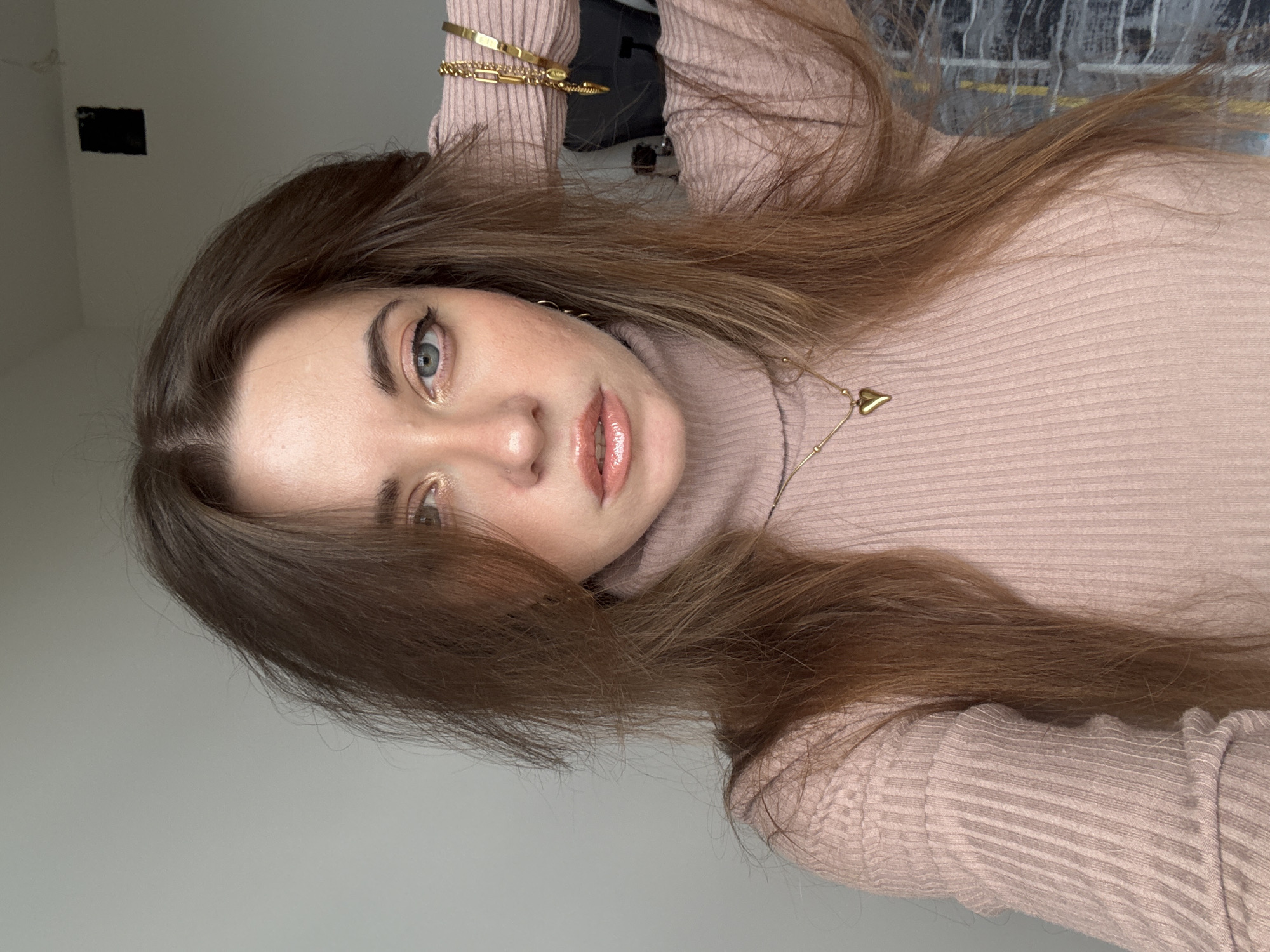 A woman with long brown hair and blue eyes is wearing a pink turtleneck sweater and gold jewelry. She is holding her hand up to her hair. The background includes a wall and part of a window. The location is not discernible.