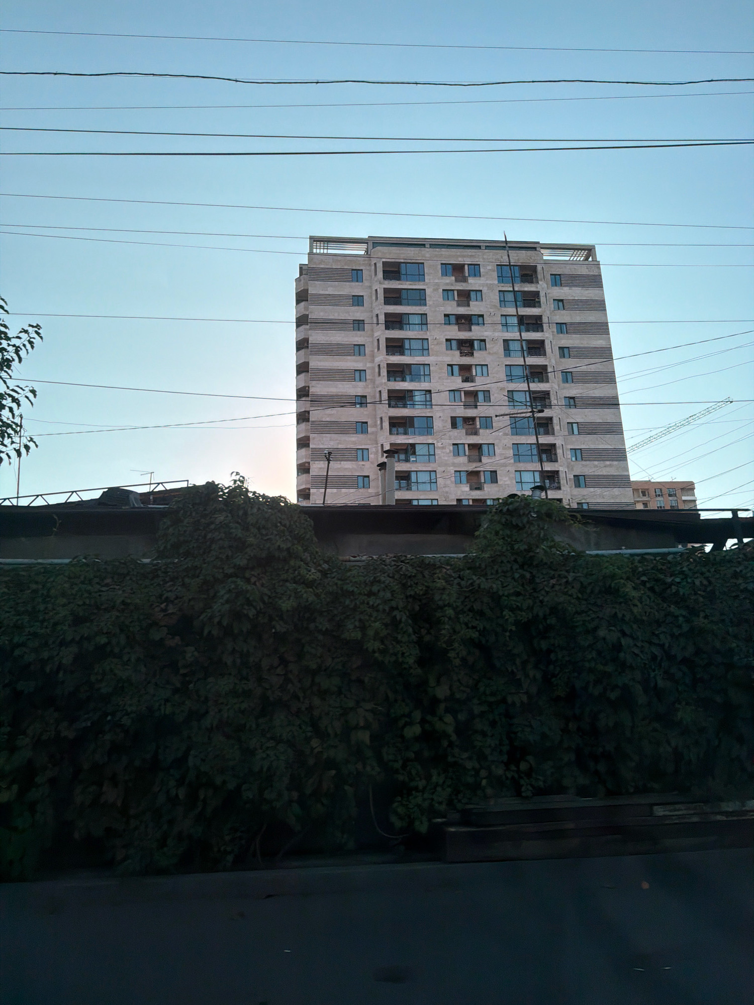 The image captures a view of a tall, modern apartment building in Yerevan, Armenia. The building, constructed with beige and grey horizontal bands and numerous windows, stands against a clear blue sky. In the foreground, a dense, dark green hedge covers a low wall. Overhead, several power lines crisscross the frame. To the right of the main building, a partially visible construction crane and another, shorter building can be seen, suggesting ongoing development in the area. The lower portion of the image shows a dark asphalt surface, likely a road or sidewalk.