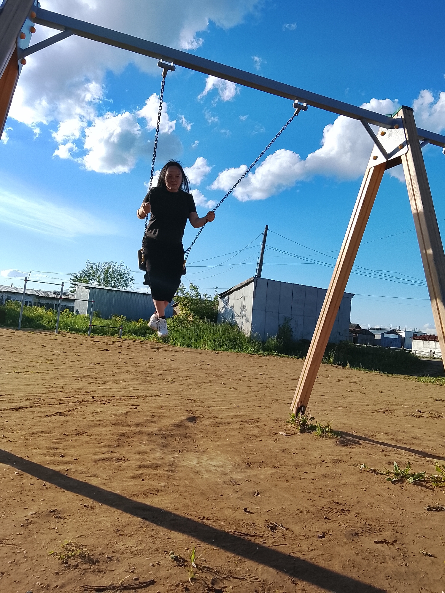 A woman is swinging on a playground swing set in what appears to be Kuyeda, Russia. The sky is bright blue with scattered white clouds. The ground is mostly dirt. In the background are various sheds and structures typical of a rural Russian setting. The woman is wearing a black outfit and white sneakers.