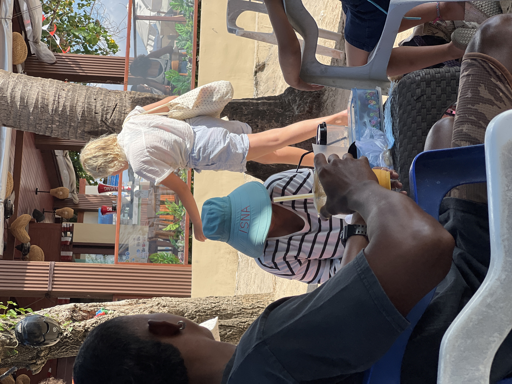 This image depicts an outdoor scene in Kuta, Indonesia, likely near a beach or coastal area. In the foreground, a person's dark-skinned arm and torso are visible, with the arm resting on a blue plastic chair. This person appears to be holding a cup with a yellow beverage and a straw, suggesting they are drinking. To the left, another person's dark-skinned shoulder and neck are visible, wearing a dark t-shirt and a silver hoop earring.
In the middle ground, a person with blonde hair, wearing a white short-sleeved shirt and light blue striped shorts, walks away from the viewer. They are carrying a patterned tote bag over their shoulder. Another person is seated, wearing a striped shirt and a wide-brimmed light blue hat with red lettering that reads "ISNA". They are also holding a cup.
The background reveals a structure with wooden pillars and a thatched roof, possibly a cafe or bar. There are tables and chairs, and some patrons are visible in the distance. Beach umbrellas and sand are also present, reinforcing the coastal setting. The overall atmosphere is casual and appears to be a sunny day.