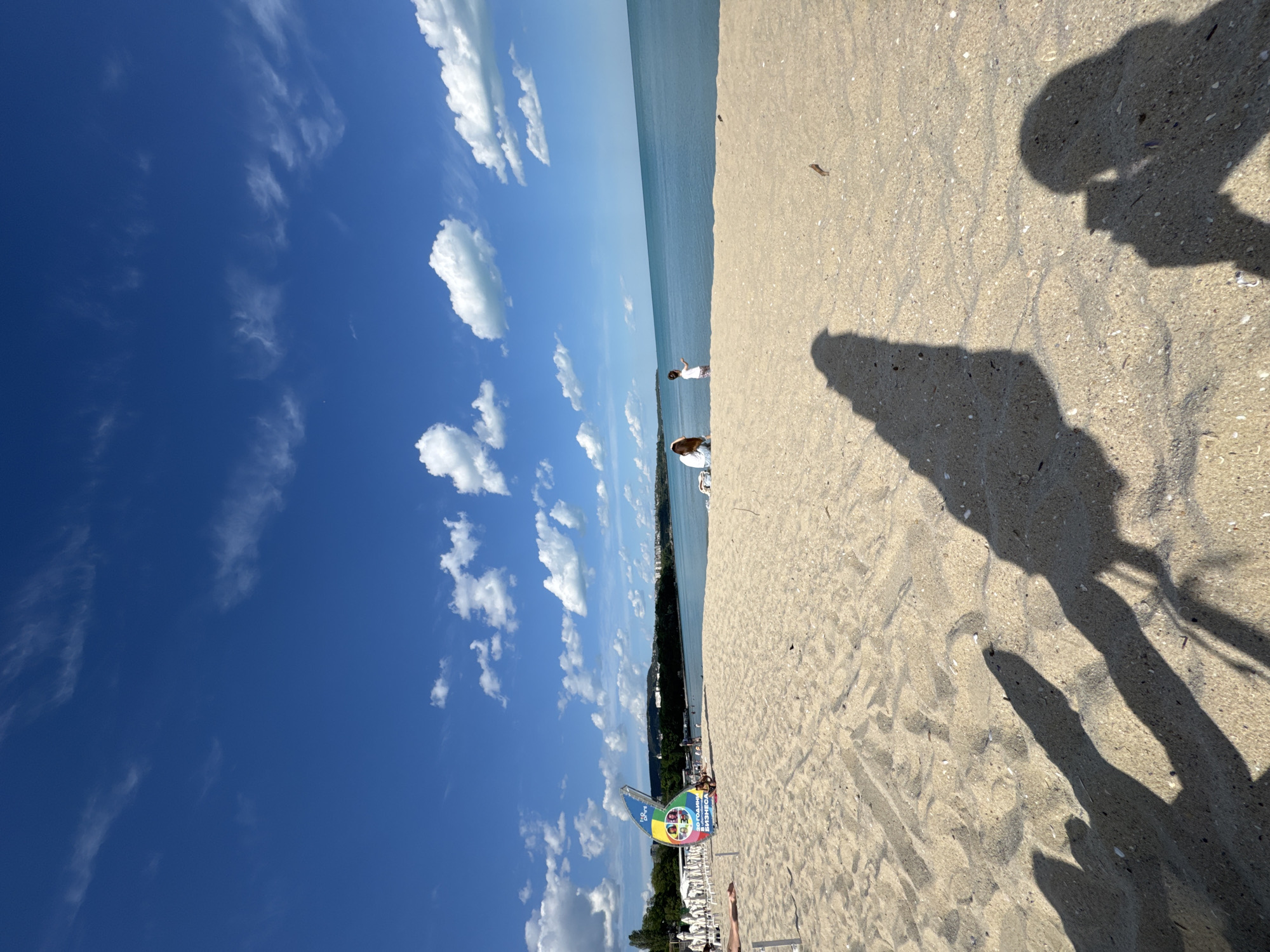 The image captures a sunny day at the beach in Varna, Bulgaria. The sky is bright blue with scattered white clouds. In the distance, the sea stretches out to the horizon. The sandy beach occupies the right side of the frame. A person and woman in white are visible towards the sea. The foreground of the beach shows the shadows of two people, possibly taking the photo, and of a sign that reads "150 години БИЗНЕС". The background shows trees and some buildings.