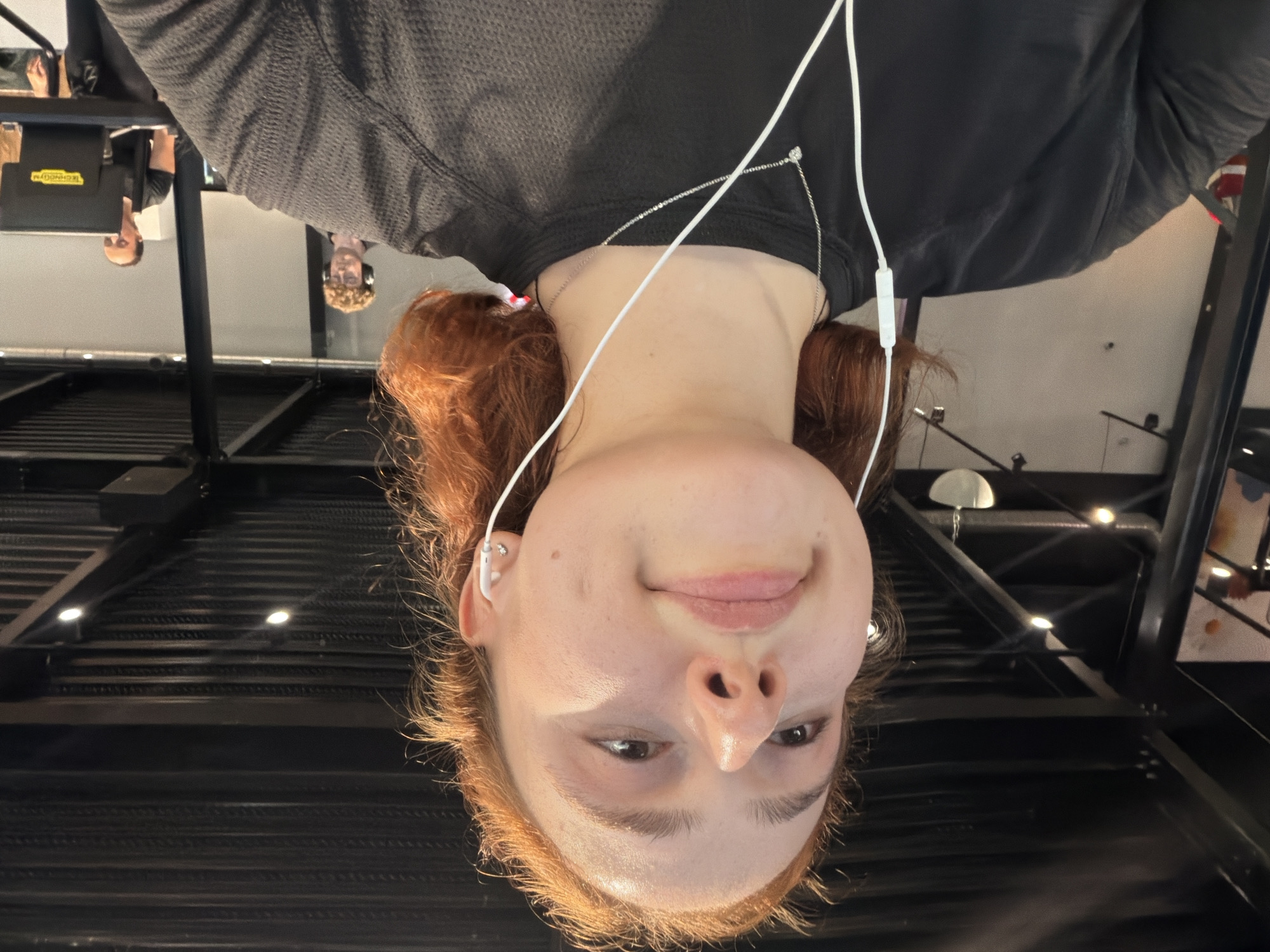 An indoor, overhead shot captures a young person from the chest up, looking directly towards the camera with a subtle smile. They have light skin, reddish-brown hair that frames their face, and are wearing a black long-sleeved top. White wired earphones are in their ears, with the wire running down their chest where a delicate silver chain necklace with a small pendant is also visible.
The background, which appears to be a reflective surface such as a mirrored ceiling or polished floor, shows reflections of the environment and other individuals. In these reflections, one person with headphones is visible, and another person is seen near gym equipment, which has a yellow "TECHNOGYM" logo displayed on it. The setting features industrial-style ceilings with dark beams and several light fixtures.
The main subject appears to be either lying down and looking up or being photographed from a high angle. Their expression is calm and pleasant. The reflections suggest the scene takes place within a modern fitness center or gym, possibly in Strasbourg, France, as indicated by the location context. The lighting is bright and even, highlighting the subject's features.