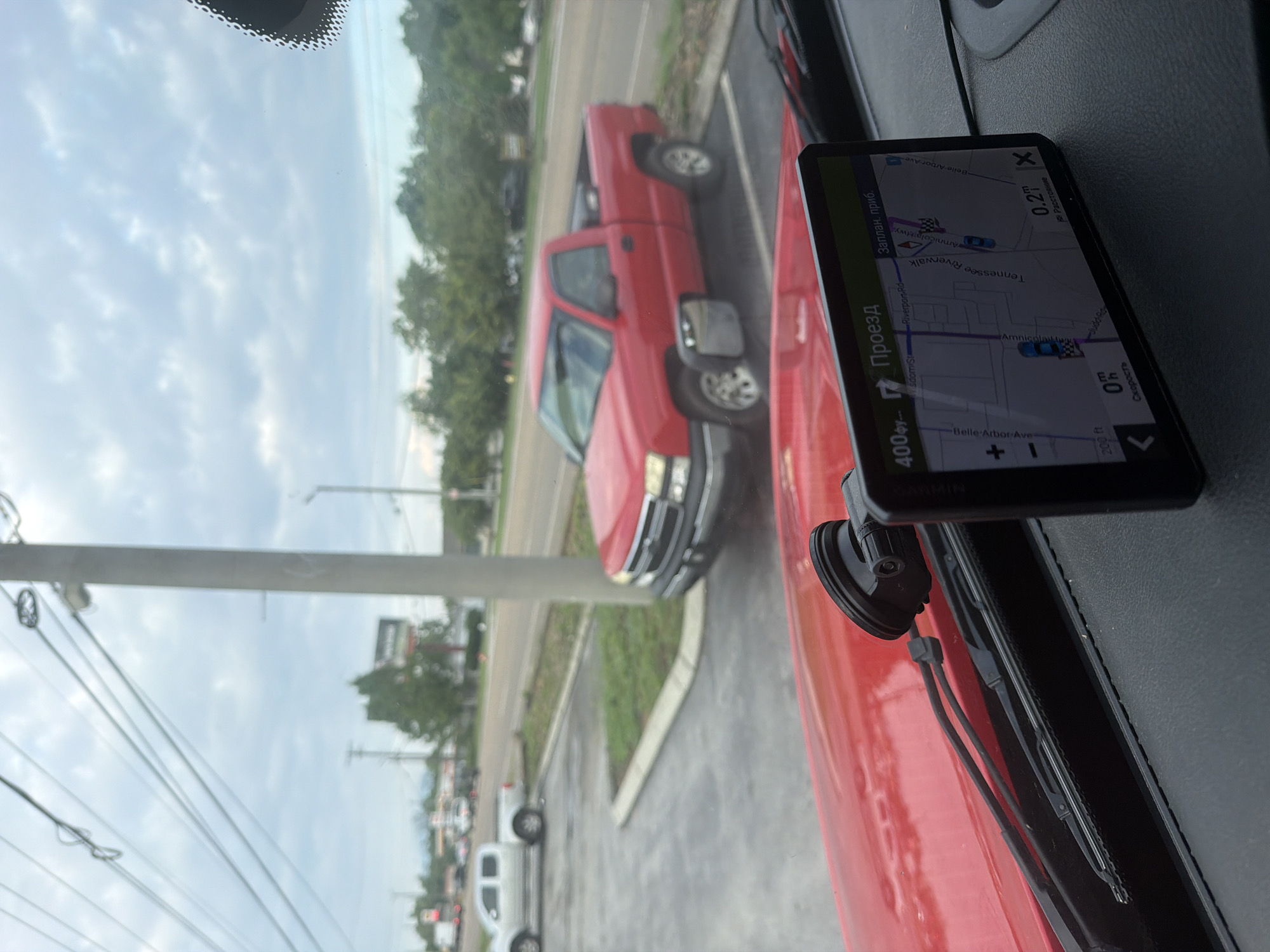 An eye-level medium shot captures the interior of a vehicle looking out a windshield at a red pickup truck parked in a lot. The pickup truck, a Chevrolet Silverado, is parked diagonally and facing left in the frame, with its driver's side door open and a side mirror extended. In the background, a white pickup truck is parked parallel to the curb on the left. A grey pole stands in the foreground between the camera and the red pickup truck, obscuring parts of the view. Beyond the parking lot, a road is visible with cars driving on it, and trees line the horizon under a cloudy sky. A GPS device is mounted on the vehicle's dashboard, displaying a map with navigation information. The image appears to have been taken in Chattanooga, Tennessee, given the displayed road names on the GPS such as "Tennessee Riverwalk".