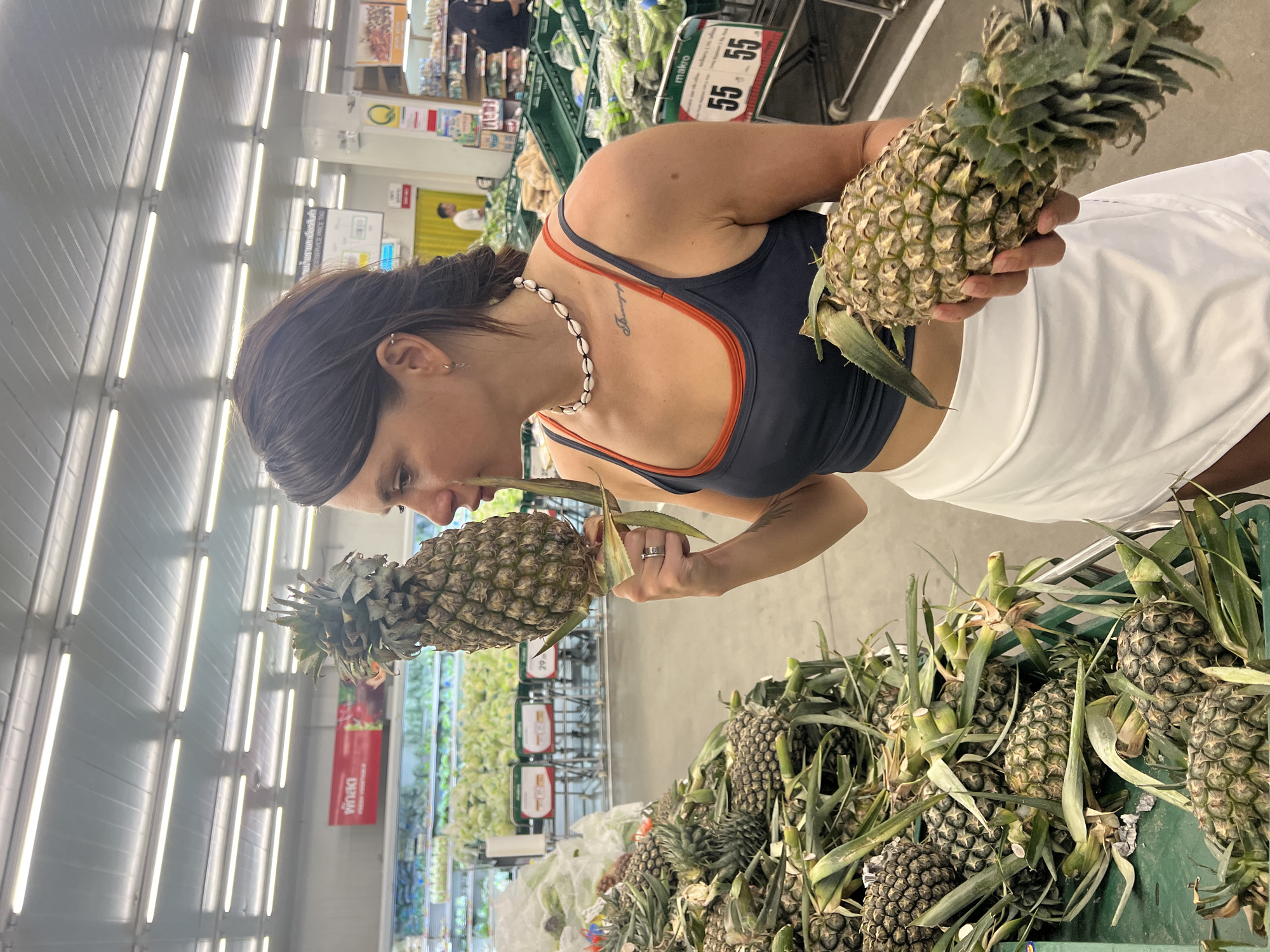 In a brightly lit grocery store in Baan Bo Phut, Thailand, a woman with dark hair, wearing a dark blue tank top with orange trim, a white tennis skirt, and a shell necklace, stands facing a display of pineapples. She is holding two pineapples, one in each hand, and appears to be examining them closely. Below her is a green plastic container filled with many more pineapples. In the background are shelves of produce, price signs, and other shoppers. The scene is inside a typical grocery store setting.