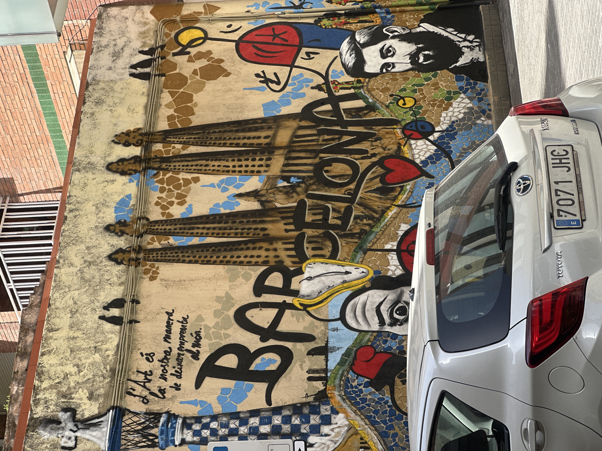 The image depicts a section of a vibrant mural on a building wall in Barcelona, Spain. The mural prominently features the word "BARCELONA" in large, stylized black lettering. Behind the lettering, a depiction of the Sagrada Familia basilica, an iconic landmark of the city, is visible with its distinctive towers. To the left of the word "BARCELONA," a surrealist melting clock, reminiscent of Salvador Dalí's work, is integrated into the artwork. The mural also includes portraits of individuals, one with a beard and another whose eyes are prominent. Abstract colorful elements and mosaics are scattered throughout the mural, contributing to its dynamic and artistic nature. The mural is painted on a textured, light-colored wall, likely part of an urban building. In the foreground, the rear of a white Toyota Yaris hybrid car is visible, partially obscuring the lower portion of the mural. The car's license plate is partially visible, showing "7071 JHC". The environment appears to be a sunny day in an urban setting, with a hint of a brick building and white railings in the upper background.