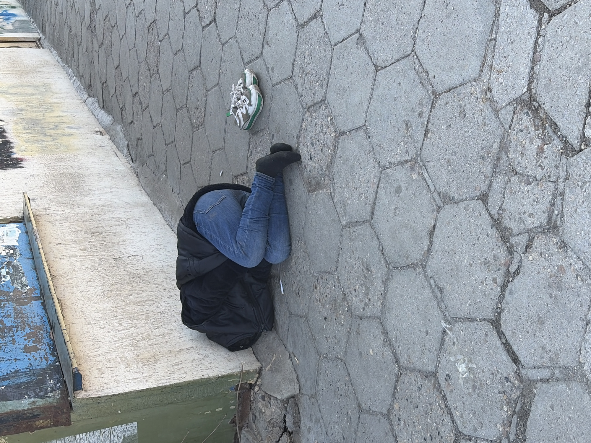 A person in blue jeans and black socks is bent over, head facing down, with a black jacket around their upper body and head. A white and green sneaker lies on the ground near the person's feet. The person is located on a stone-tiled walkway beside a concrete ledge, which is adjacent to a blue and gray structure. The background consists of a wall made of hexagonal stone tiles. The location is outdoors. The location is presumed to be in Sofia, Bulgaria.