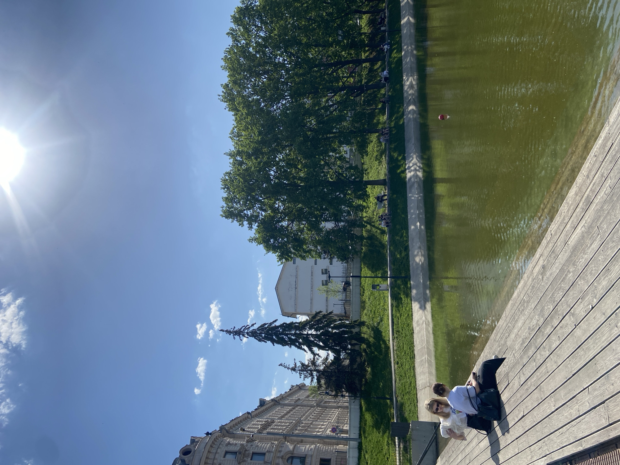 The image shows a view in Kazan, Russia, likely near a body of water. A pond or canal stretches across the frame, and in its waters floats a red ball. A wooden deck or boardwalk runs alongside the water's edge where two young women are sitting. One wears a white top and the other is in a white top and sunglasses. They are looking at a phone. Beyond the pond, a grassy area borders a sidewalk lined with trees and several individuals are sitting along it. A large building is visible in the distance, and the sky is blue with scattered clouds and a bright sun. A tall conifer tree stands prominently among other trees in the cityscape.