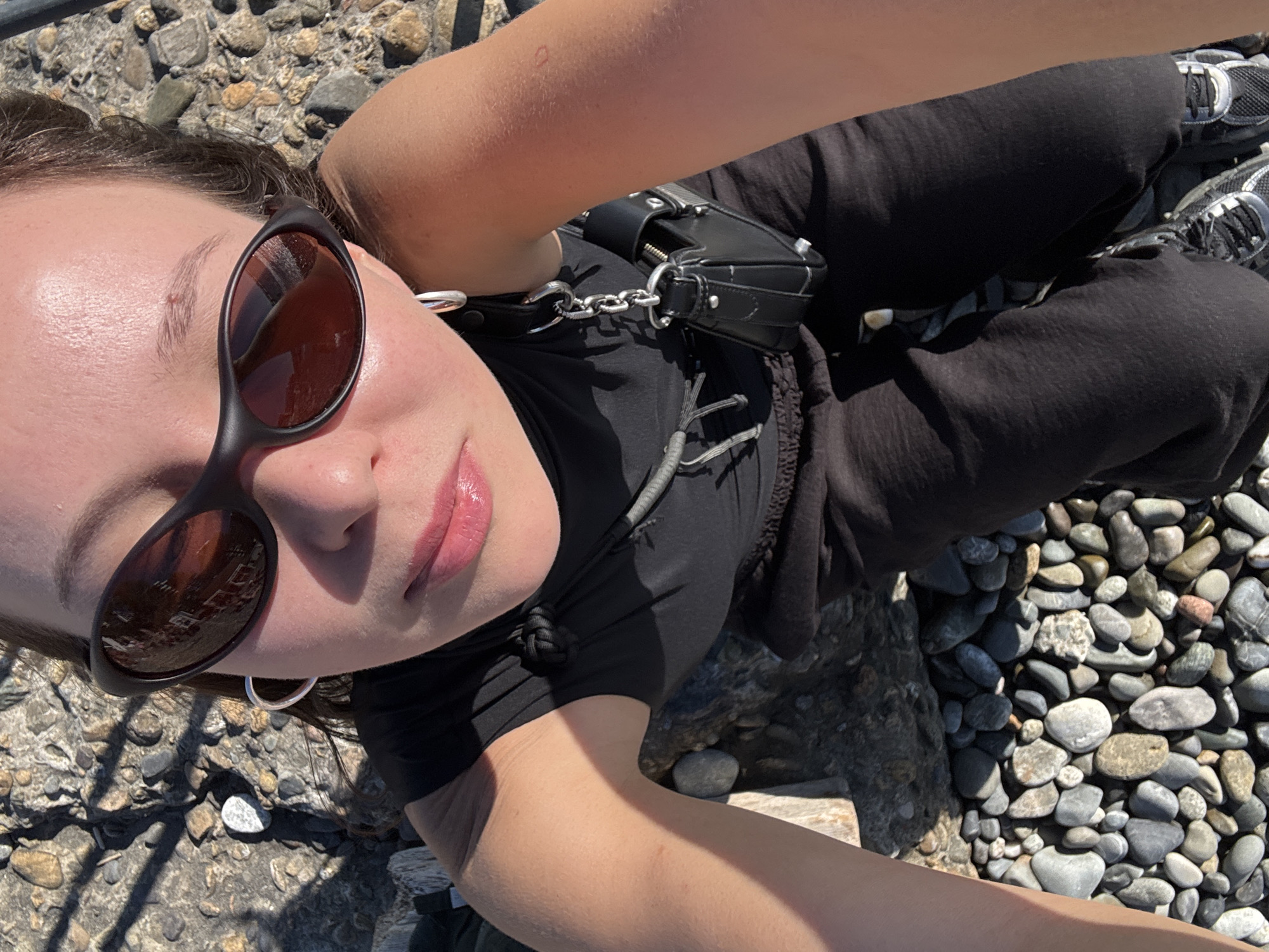 A woman is outdoors on a sunny day, positioned on a rocky surface, possibly a beach or embankment, in Sochi, Russia. She is looking up at the camera, wearing black sunglasses, a black sleeveless top, and black pants. A small black purse is attached to her clothing. Her skin is illuminated by the sun, creating shadows on the ground and her body. The background consists of numerous smooth, grey and brown stones, with some larger rocks and driftwood also present.