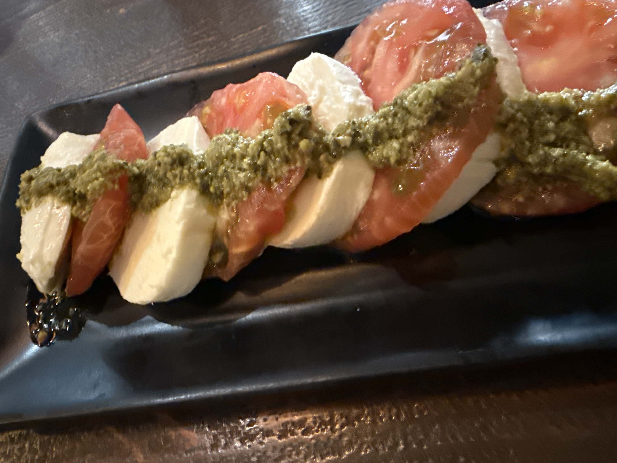 A Caprese salad is presented diagonally on a black, rectangular plate with raised edges. The salad consists of alternating slices of white mozzarella cheese and red ripe tomatoes, stacked vertically. A thick, textured green pesto sauce is drizzled in a zig-zag pattern over the entire stack. A small pool of dark, viscous liquid, likely balsamic glaze, is visible at the upper left corner of the plate. The plate rests on a dark, textured surface, possibly wood, with an area to the left showing a more distinct rough texture and reflections. No individuals are present. The scene is located in Unknown, Unknown.