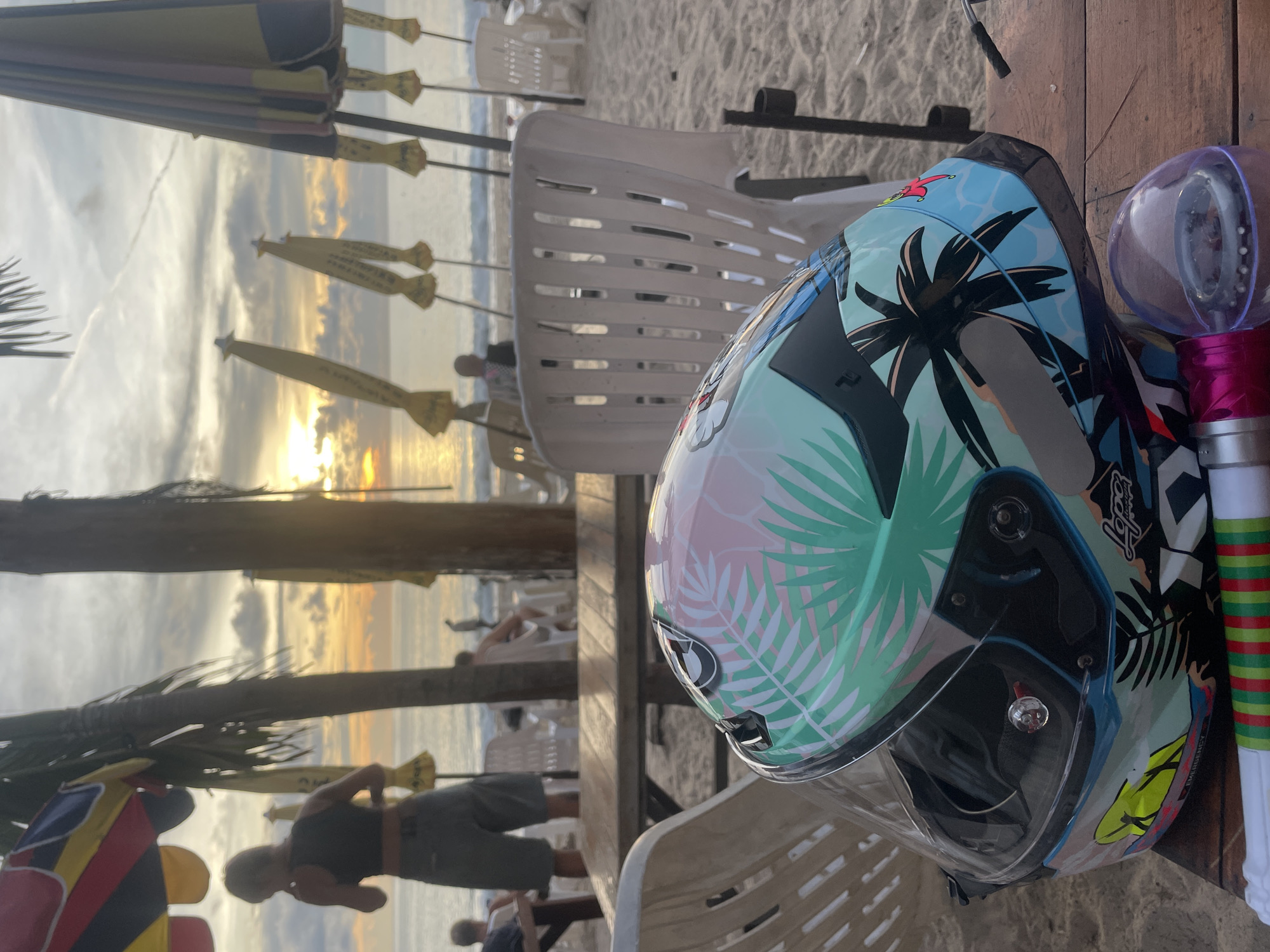 The image shows a motorcycle helmet and a purple light stick on a wooden table at a beach in Choeng Thale, Thailand. The helmet has a colorful design with palm trees and leaves. White plastic chairs are visible around the table. In the background, there is a beach with umbrellas and people, with a sunset in the distance. The image is taken during daylight.