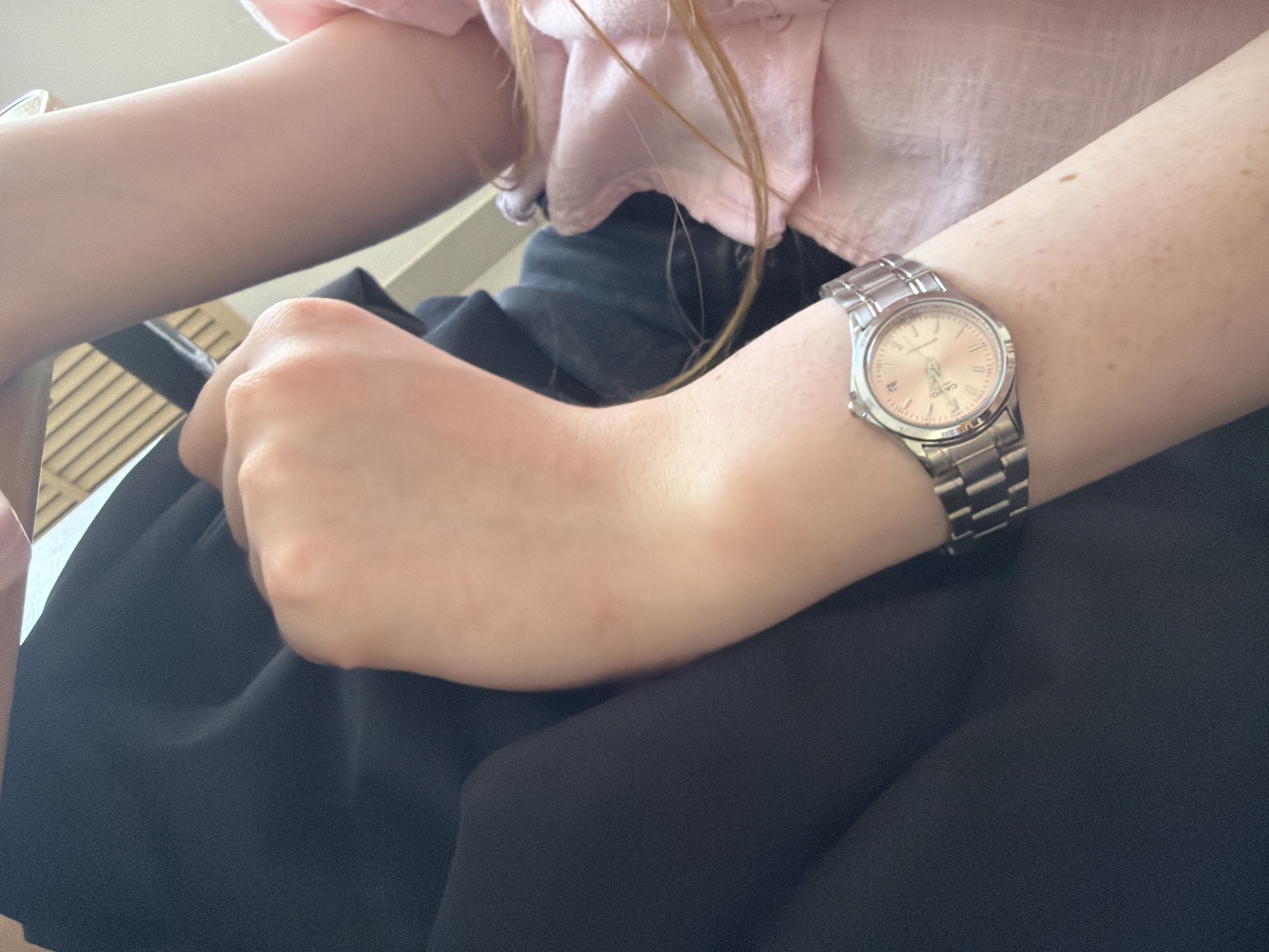 A person wearing a pink ruffled blouse and a black skirt sits with their left arm resting on their leg. A silver-toned wristwatch is on the person's left wrist. The scene appears to be indoors, near a wicker or woven chair. There is no information visible in the image that indicates the location is in the city of Chaikovsky, Russia.