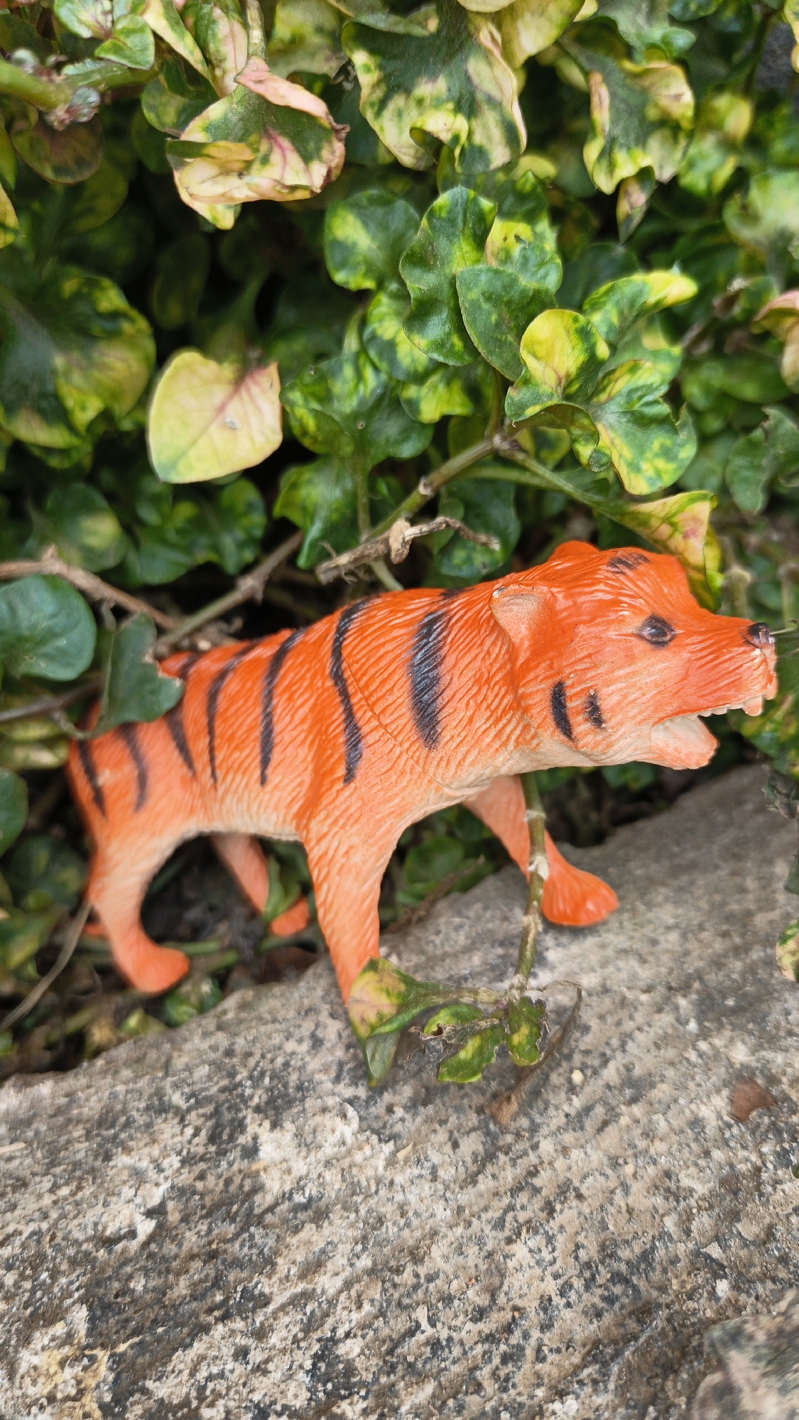 A small, orange plastic tiger toy with black stripes stands on a rough, grey stone surface. It is positioned amidst green and yellow variegated leaves, with some brown twigs visible. The background consists of dense foliage. The scene appears to be outdoors, possibly in a garden or natural area in Desa Arjasari, Indonesia. There are no people present, and the tiger is inanimate.