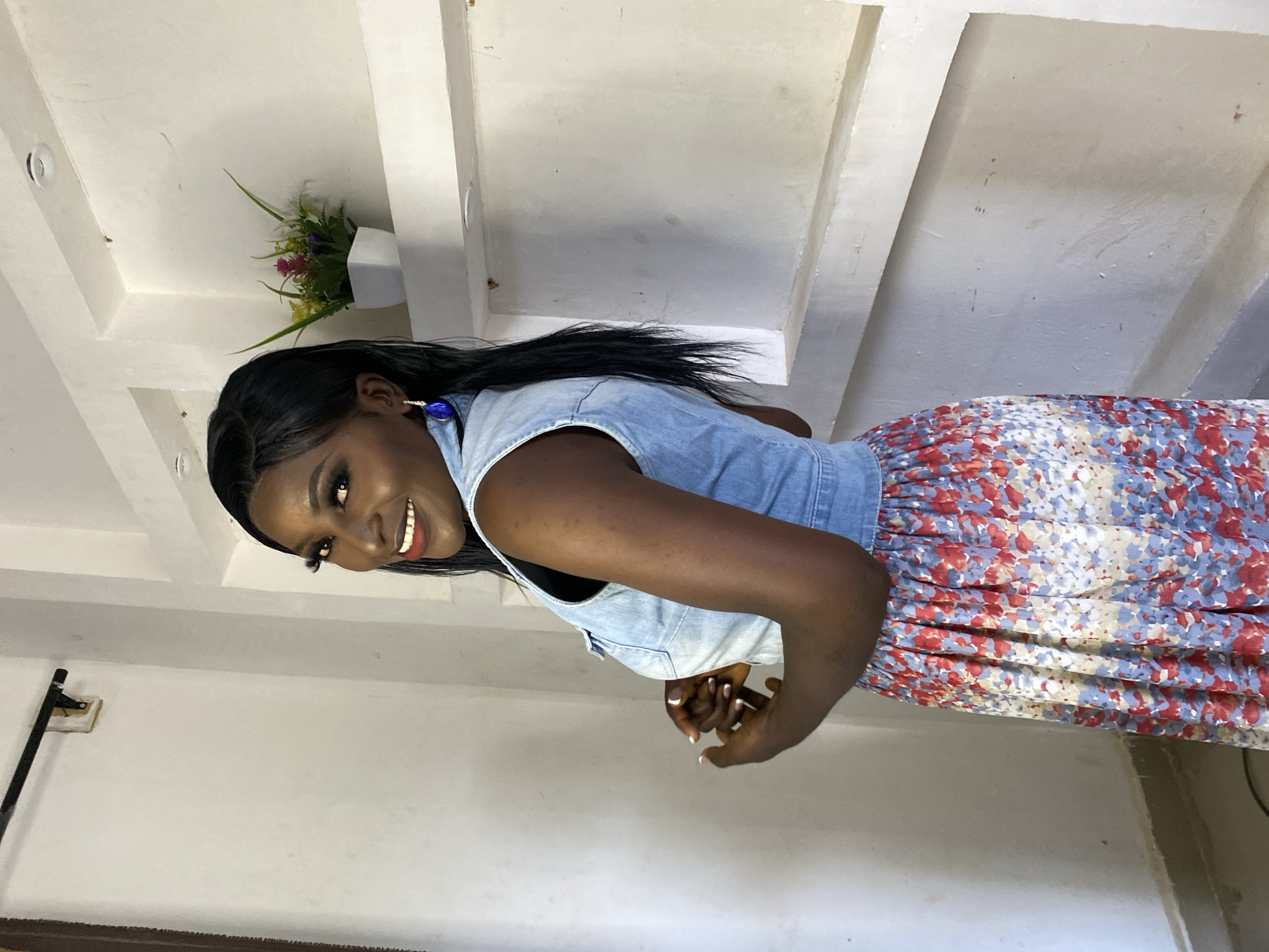 A medium shot shows a Black woman standing in front of a textured white wall with recessed shelves. She is wearing a light blue denim vest over a black top and a long, floral-print skirt with red, blue, and cream colors. Her dark hair is styled long and straight. She wears large blue teardrop earrings and has on dramatic makeup, including long false eyelashes. The woman is looking over her left shoulder at the camera with a smile. In one of the shelves above her, there is a small white pot with green and pink artificial flowers.