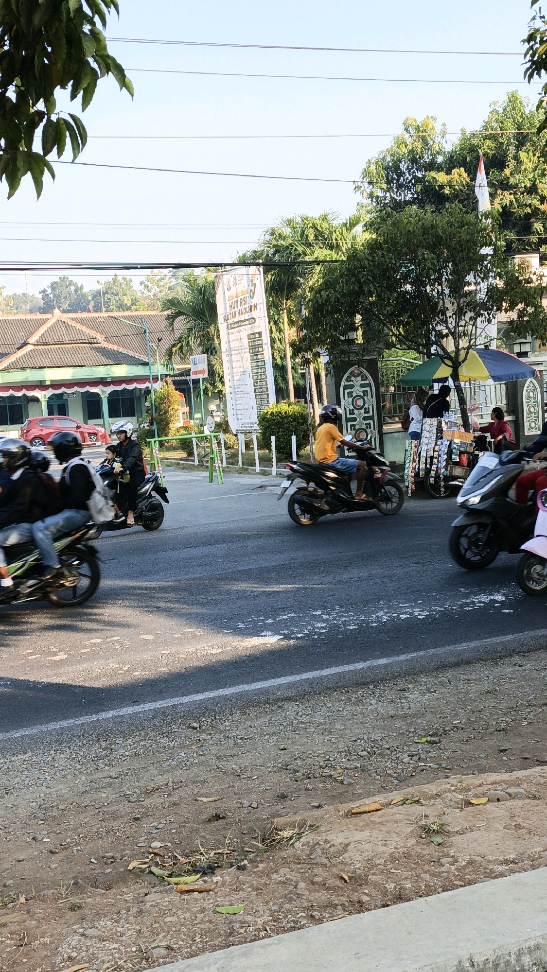 The image depicts a street scene in Kuwasen, Indonesia, on a bright, clear day. In the foreground, a paved road is visible, with a gravel shoulder to the right. Several motorcycles are in motion, indicating traffic flow. On the left side of the road, a red car is parked. Behind the road, there is a building with a red tiled roof and green trim, possibly a public building or a shop. Further in the background, a banner with Indonesian text, including "HUT RS SULTAN HADLIRIN," is displayed, suggesting a celebration related to a hospital. Trees and greenery are abundant throughout the scene, and a small stall with colorful umbrellas is set up near the entrance of a gate. People are seen interacting at the stall, and some are riding motorcycles. The overall atmosphere is that of a typical day in a bustling Indonesian town.