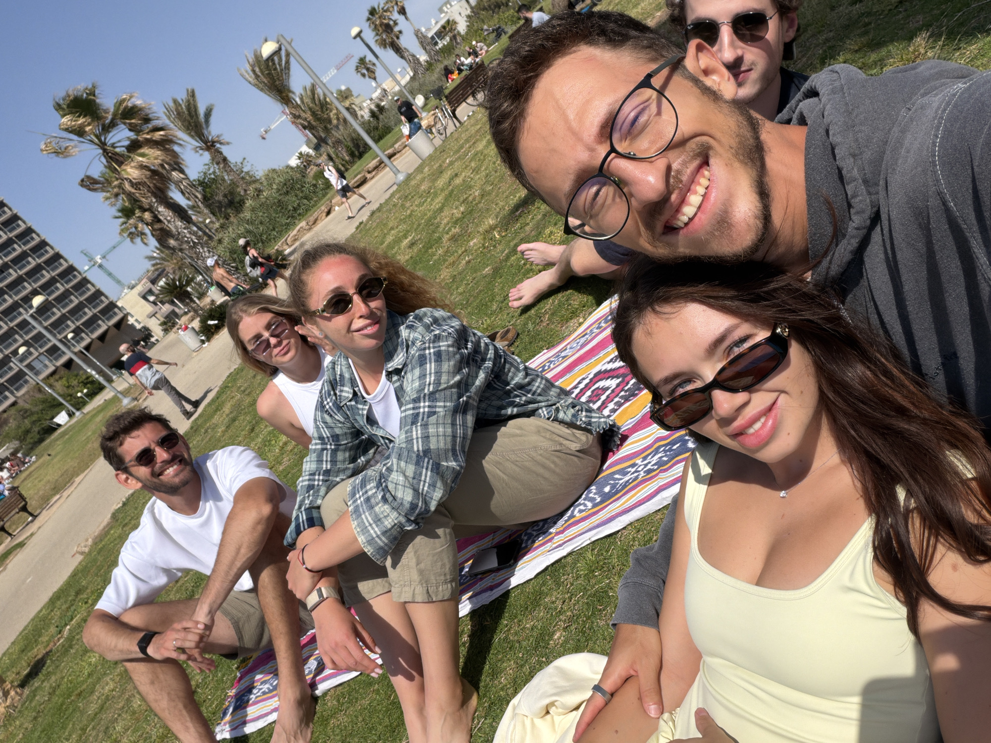 The image captures a group of six people posing for a photo on a sunny day. The setting appears to be a park in Tel Aviv, Israel, given the presence of palm trees and a tall, modern apartment building in the background. The group is sitting and lying on a colorful striped blanket spread on the grass. From left to right, there is a man in a white shirt and sunglasses sitting cross-legged, followed by two women seated, one in a white tank top and the other in a green and grey plaid shirt. Behind them, there is another man in sunglasses. In the foreground, a man with glasses and a grey hoodie is holding the camera. Next to him is a woman in a yellow tank top and sunglasses. The people are smiling and seem to be enjoying the outing. The park appears to have a paved path with people strolling, benches, and other trees and vegetation. The lighting is bright and sunny.
