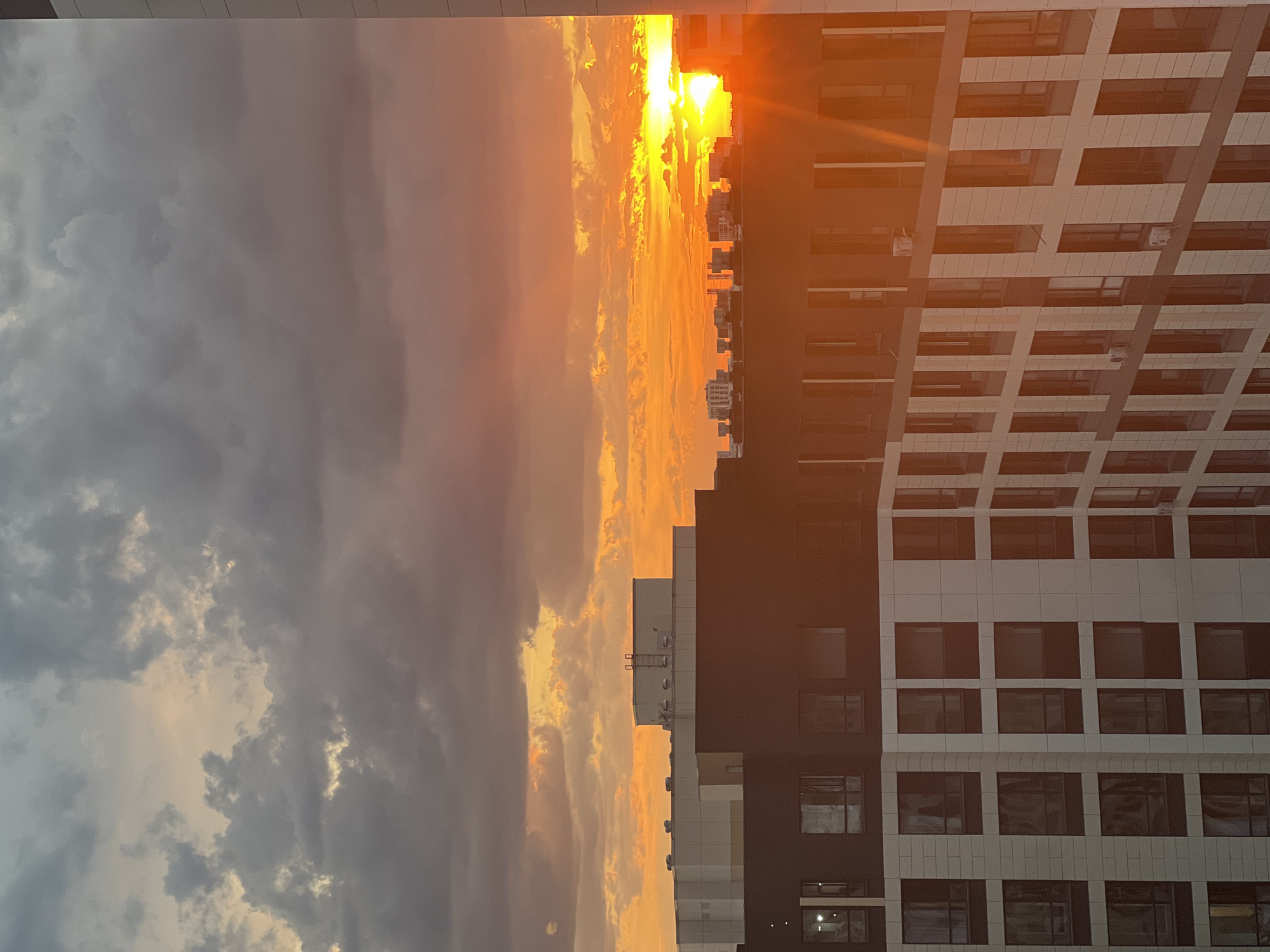 This image depicts a cityscape of Astana, Kazakhstan, at sunset. The sky is dominated by dramatic clouds, with vibrant orange and yellow hues from the setting sun breaking through. In the foreground, modern residential buildings with dark and light facades and numerous windows are visible. The architecture suggests a contemporary urban environment. The lighting indicates it is late afternoon or early evening. There are no people or identifiable actions in the scene.