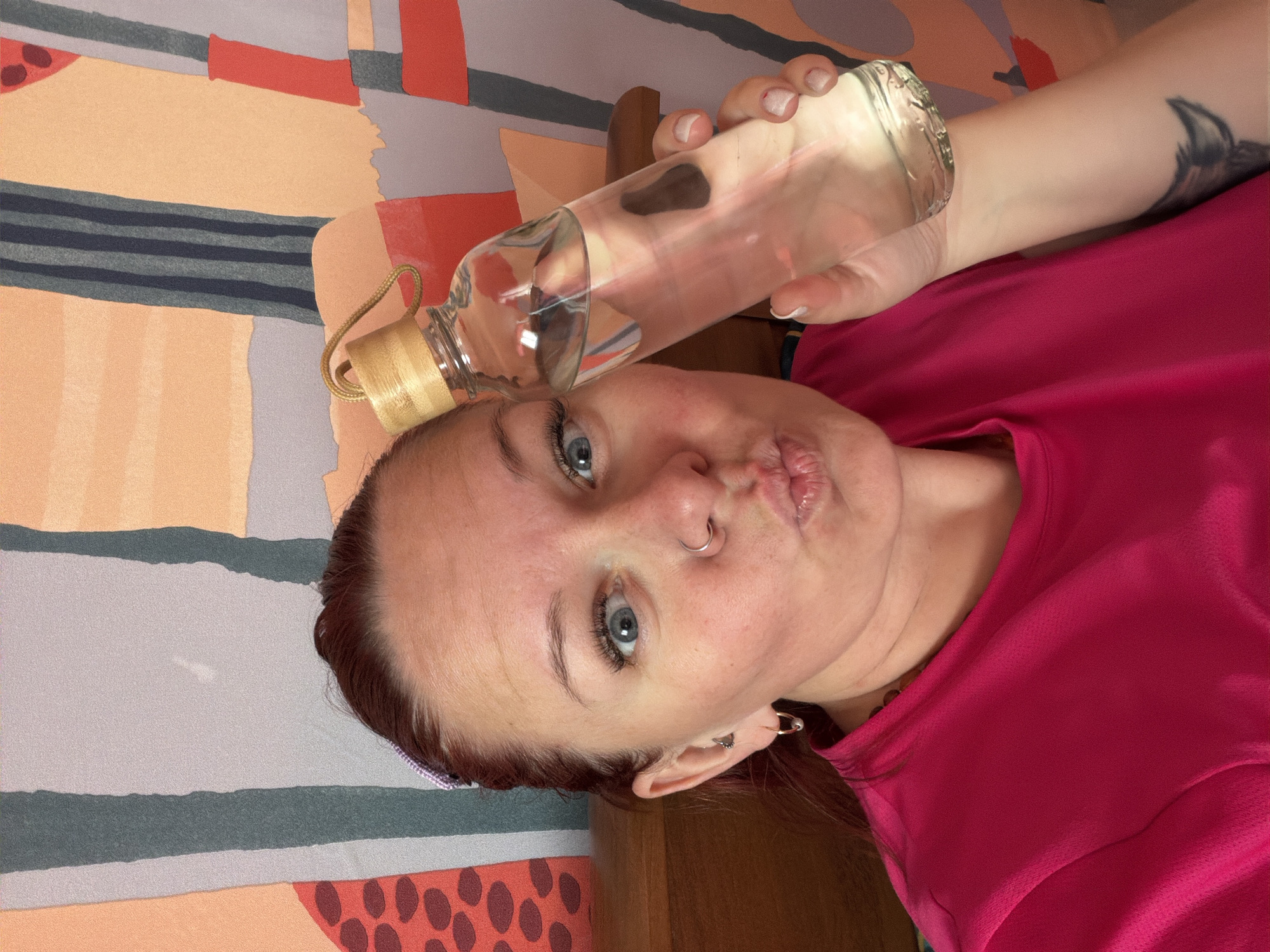 The image is a selfie of a fair-skinned woman with blue eyes and reddish-brown hair. She is wearing a pink shirt and has multiple piercings in her left ear and a nose ring. She is holding a clear glass water bottle with a wooden lid in her right hand. The background features a brightly colored patterned fabric or surface. There is no visual information in the image to confirm its location in the city of Chaikovsky, Russia.