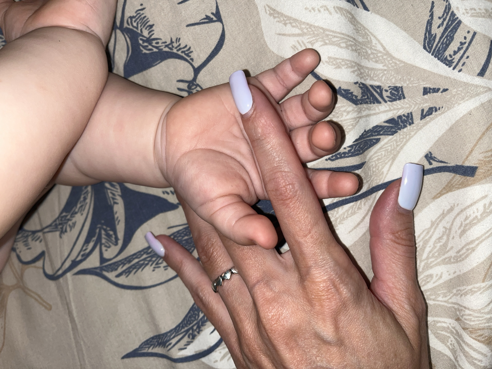 The image displays a close-up of two hands, one adult and one infant, resting on a patterned fabric surface. The adult's hand, positioned on the left, is adorned with light purple nail polish and a silver ring with a heart pattern. The infant's hand, on the right, is smaller and shows no nail polish. The fabric beneath them features a repeating pattern of stylized leaves in beige, white, and navy blue. The lighting is dim, and there are no visible landmarks to identify a specific location in Nizhny Novgorod, Russia. The image captures a moment of gentle contact between the two hands.