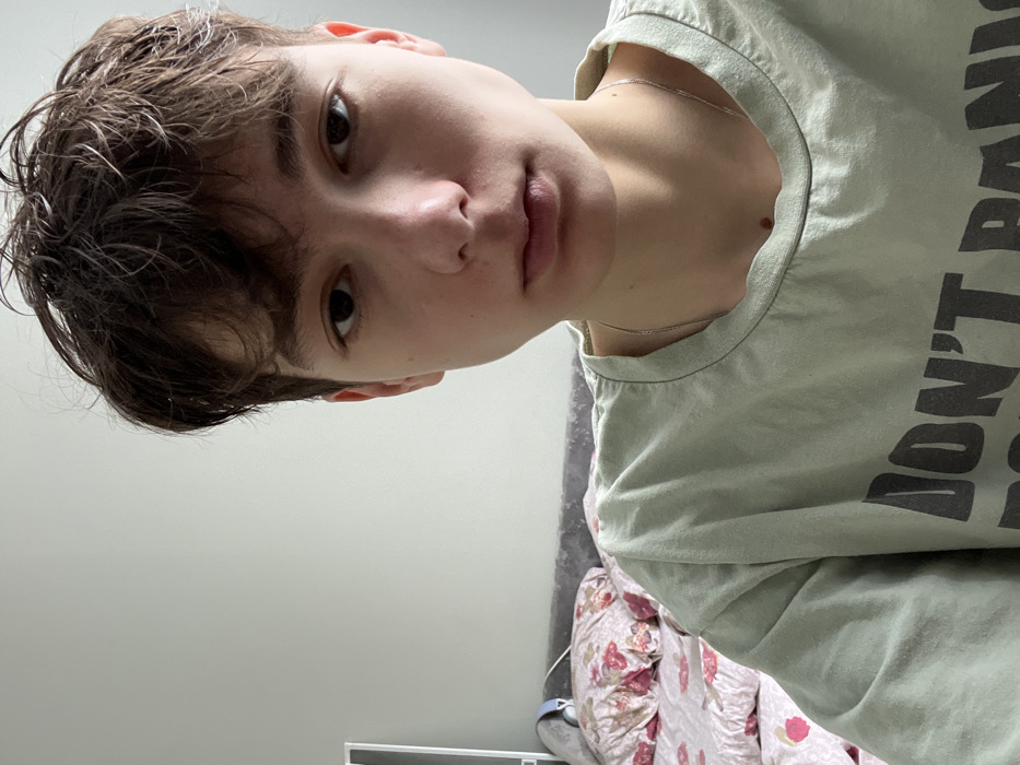 The image displays a headshot of a young individual, possibly a teenager, positioned indoors in what is identified as the city of Zainsk, Russia. The person, with light skin and short, disheveled dark brown hair, faces directly towards the camera, maintaining a neutral expression with eyes open. They are wearing a light sage green short-sleeved t-shirt featuring dark grey text, partially visible, which reads "DON'T PANIC". A thin silver chain necklace is visible around the neck.
The background consists primarily of a plain light grey wall. To the left and behind the person, a bed is visible with a dark grey upholstered headboard. The bedding includes a light-colored duvet or sheet adorned with a pink and red floral pattern. A pair of light blue over-ear headphones rests on the headboard, partially obscured, to the left of the person's head. The lighting is even, suggesting an interior light source or diffused natural light.