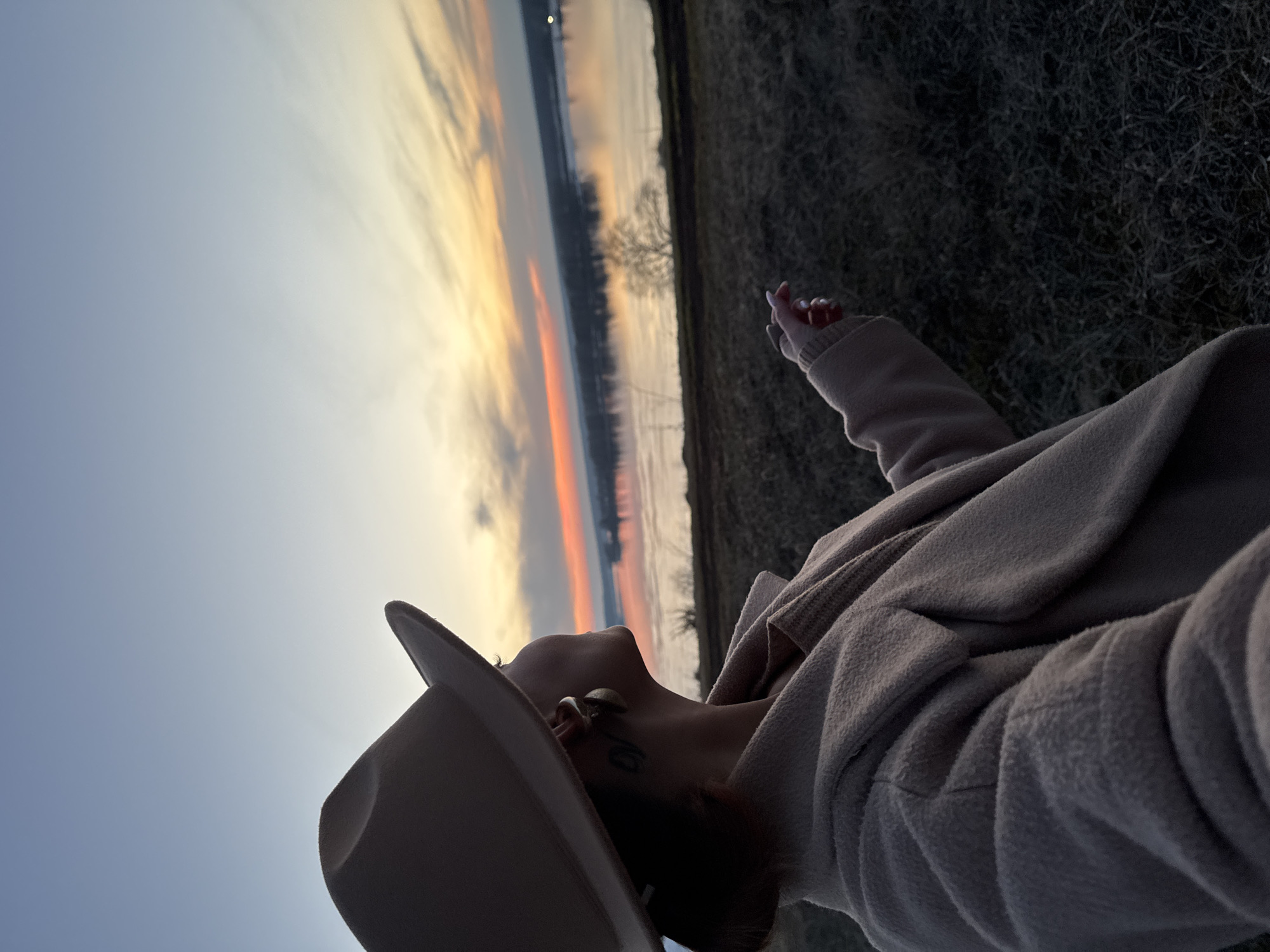 A low-angle shot of a person in Ольховка, Russia. They are wearing a wide-brimmed hat and a light-colored coat or sweater. The person is looking up and pointing a finger towards the sky. In the background, there's a sunset over water. To the right of the water is a grassy or brushy area, and some trees.