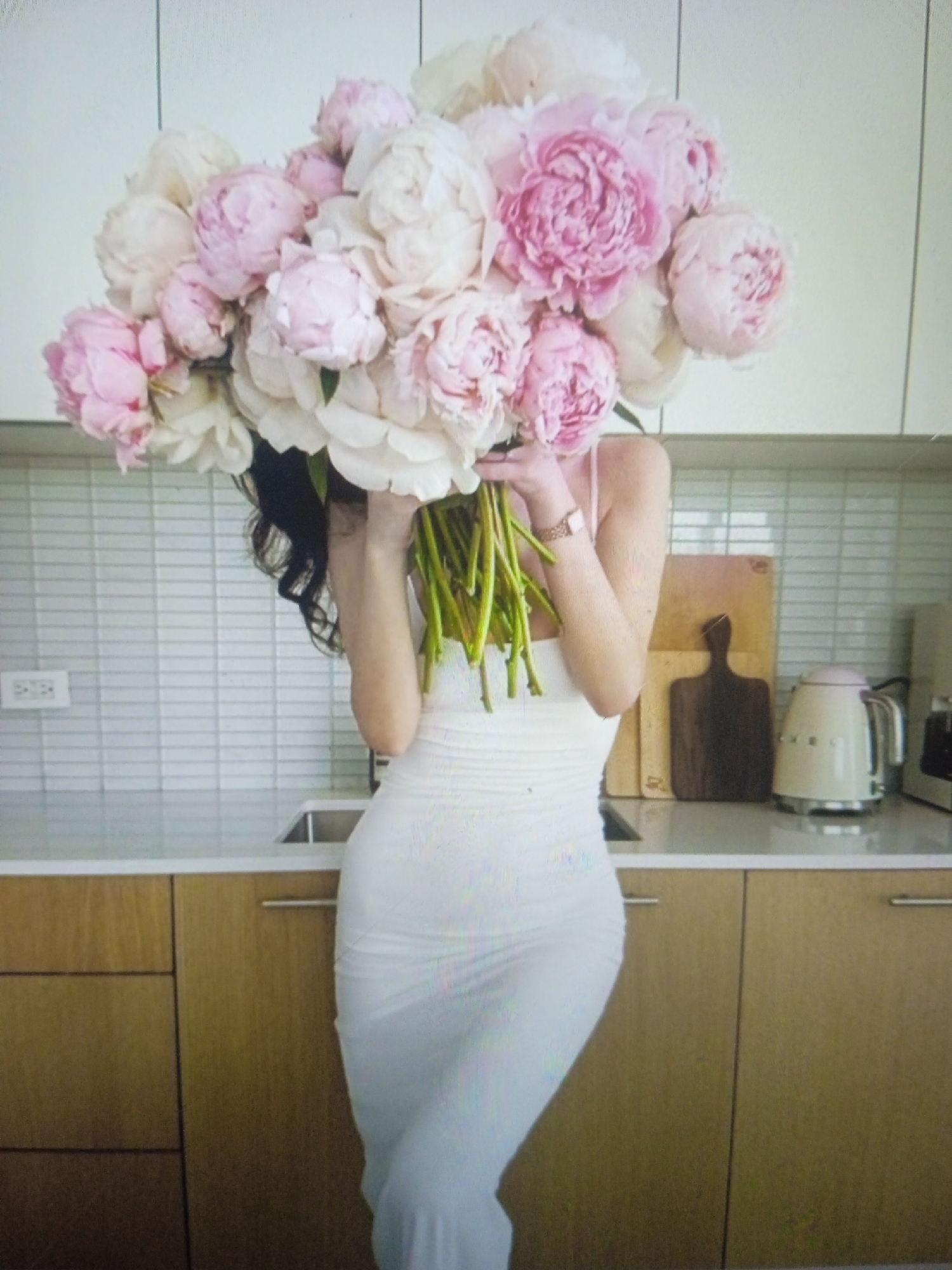 A person wearing a white bodycon dress stands in a kitchen, holding a large bouquet of pink and white peonies that obscures their face. The kitchen features light-colored tiled backsplash, a countertop with a sink and a beige electric kettle. Wooden cabinets with silver handles are visible below the countertop. The scene is brightly lit. The image is a photograph taken indoors, likely in a residential setting.