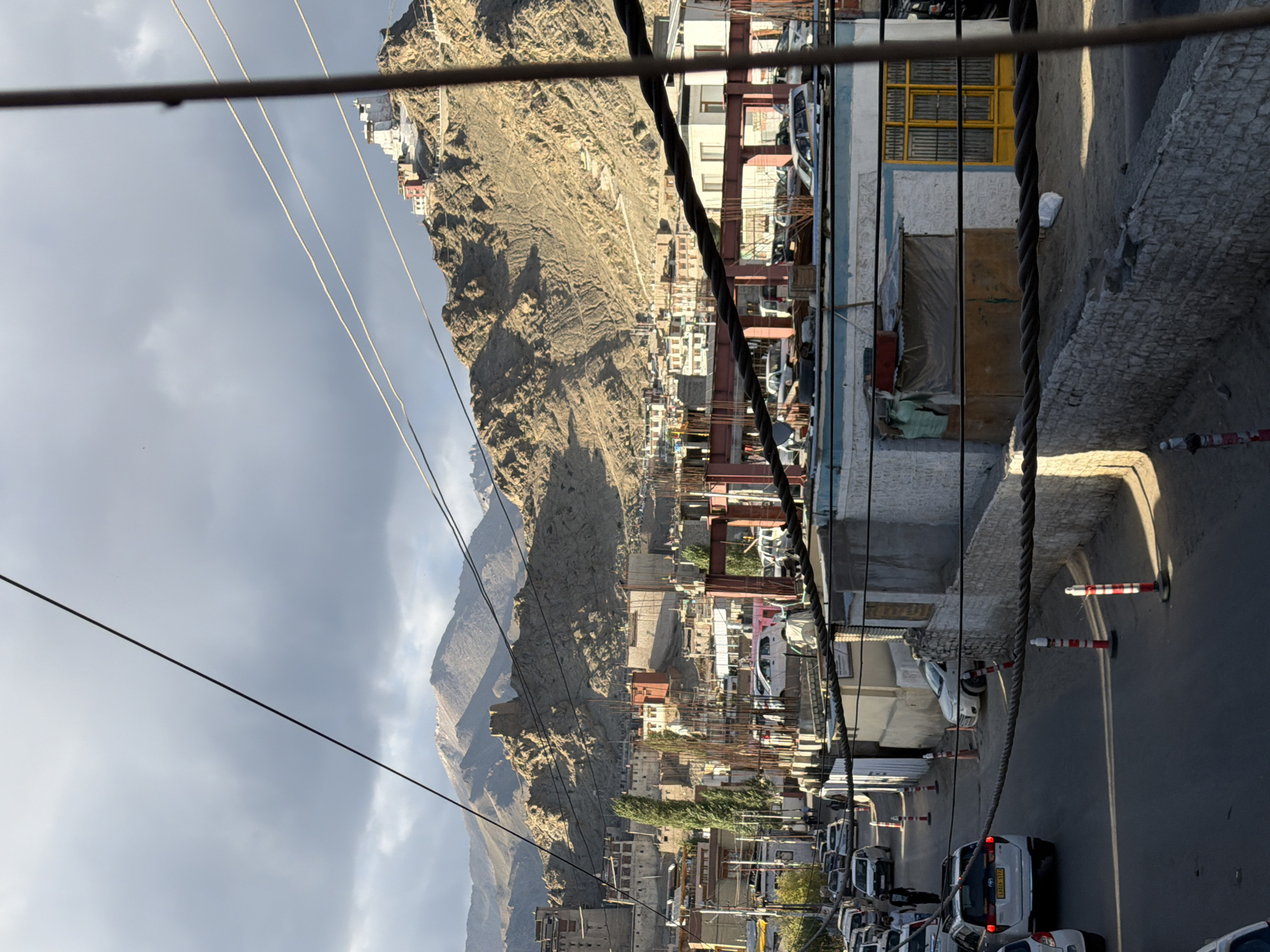The image depicts a street scene in Leh, India, with the rugged, mountainous landscape of Ladakh serving as the backdrop. In the foreground, a street is lined with parked cars and several traffic cones. Buildings with various architectural styles are visible on either side of the street, some appearing to be under construction with exposed steel framing. Power lines are strung across the scene, crossing overhead and in front of buildings. In the distance, a prominent white building with red accents sits atop a hill, suggesting a monastery or palace structure characteristic of the region. The sky is overcast with dramatic cloud formations, and the sunlight casts shadows on the mountainous terrain. The overall impression is of a bustling town nestled within a dramatic natural environment.