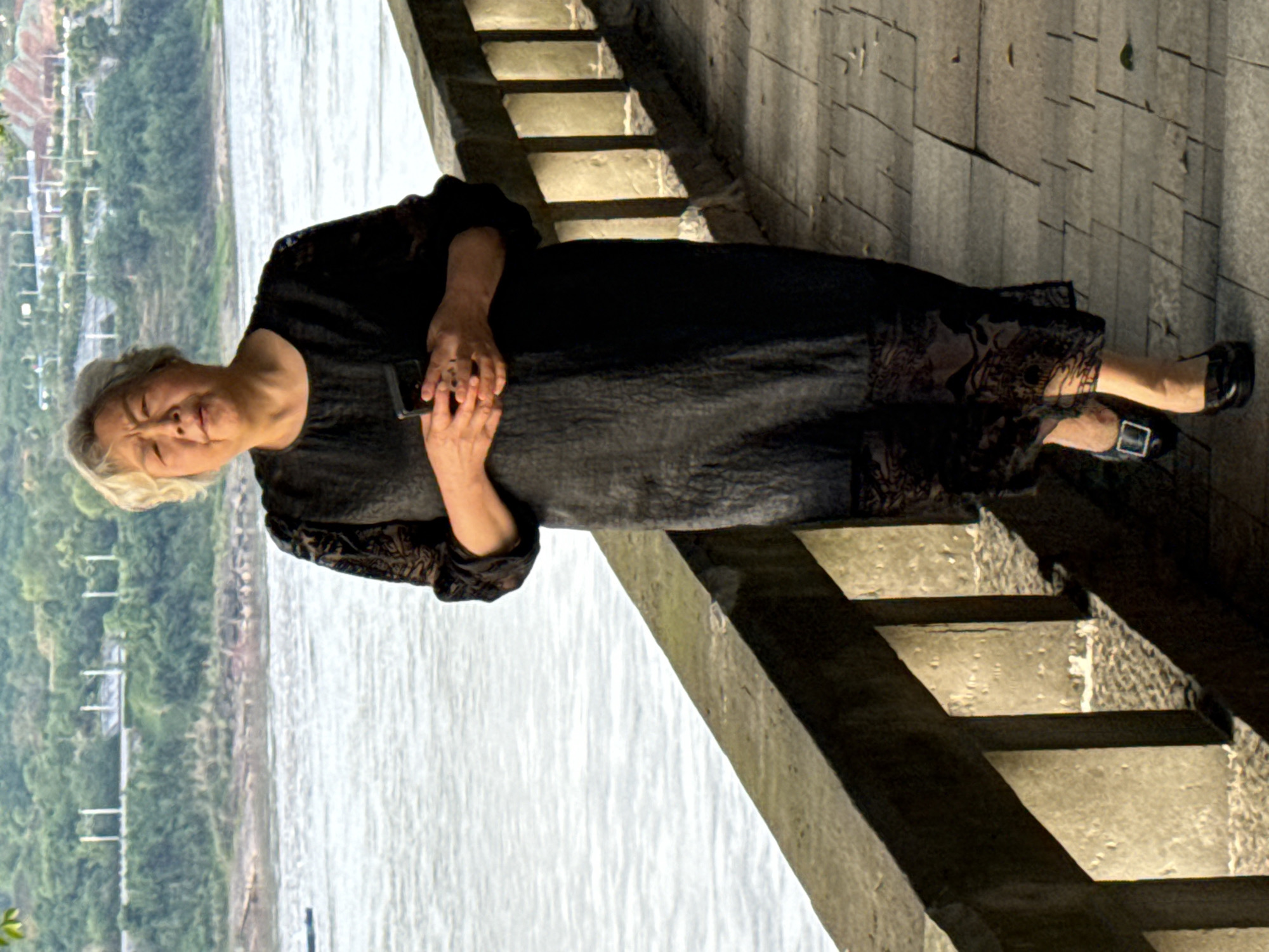 An elderly woman is depicted standing outdoors on a stone walkway or set of steps. She is dressed in a long, dark-colored gown or robe, which features subtle patterns on the sleeves and lower part of the skirt. Her hair is short and light-colored, possibly gray or blonde. With both hands clasped together, she holds a dark-colored smartphone in front of her. She wears dark flat shoes with a buckle and looks towards the viewer with a calm, gentle expression.
In the background to her left, a wide river flows, its surface showing ripples and reflections from the daylight. The distant bank across the river is covered in dense green foliage, suggesting hills or mountains, with faint outlines of what appear to be buildings or structures visible. To her right, a series of light-colored stone steps or a tiered wall is illuminated by sunlight, casting prominent shadows that indicate a clear, sunny day, likely during the morning or late afternoon. The setting is a scenic riverside area, consistent with the provided location context of Jiangjin, China. No text is visible in the image.