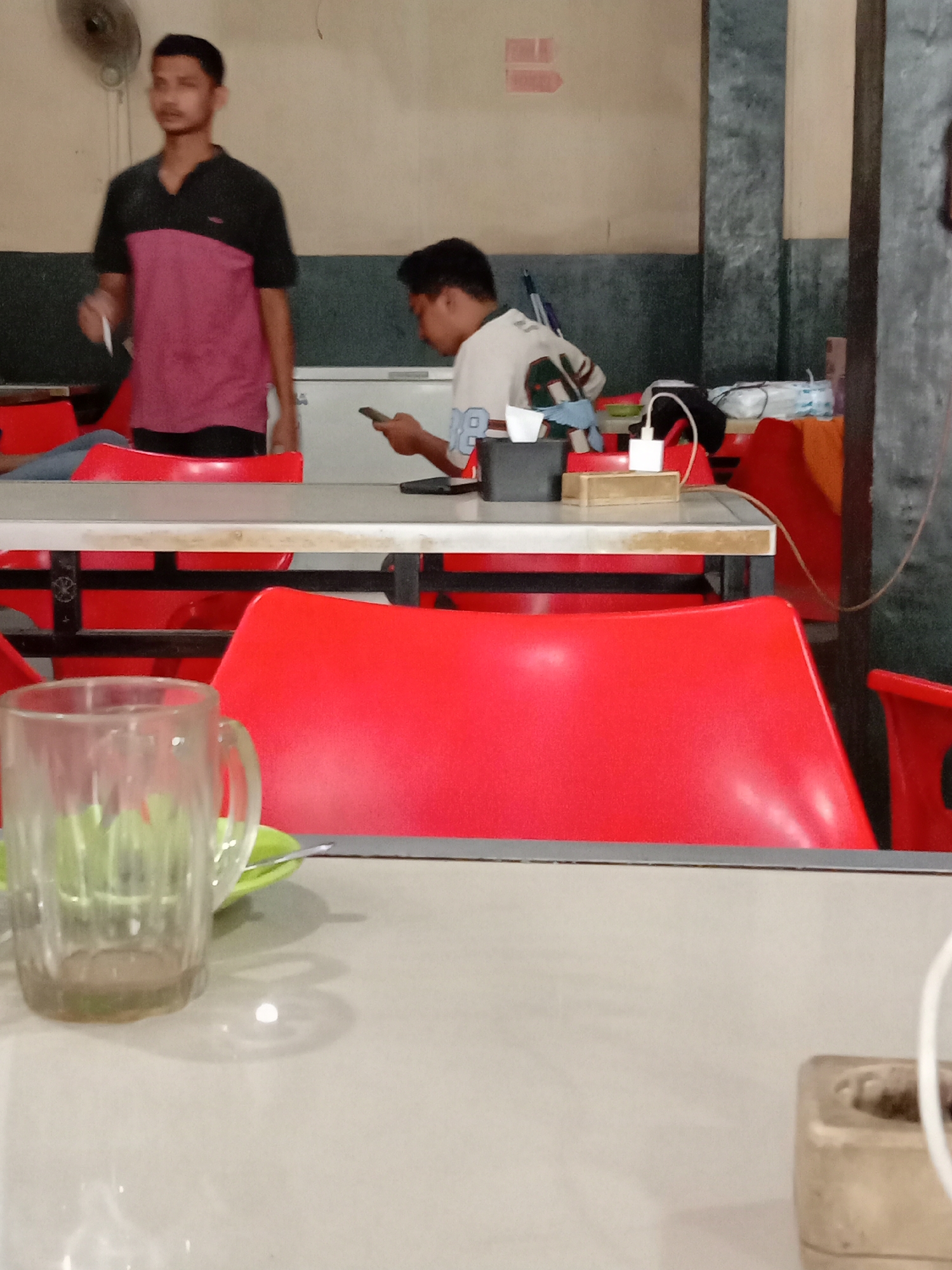 In an indoor establishment, possibly a cafe or restaurant, in Lhokseumawe, Indonesia, two young men are present. The man on the left, wearing a dark-collared shirt with a maroon panel across the chest and black shorts, stands and holds a white object, possibly a pen or cigarette. The man on the right, seated and facing away from the camera, wears a white t-shirt with dark accents and is looking at his mobile phone. He is positioned at a table with a charging cable connected to a white power adapter. The environment features red plastic chairs and tables with light-colored laminate tops. In the foreground, a glass mug with some liquid and a green saucer and spoon rest on a table. The walls are a neutral color, with a dark band running horizontally. A ceiling fan is visible in the upper left corner.