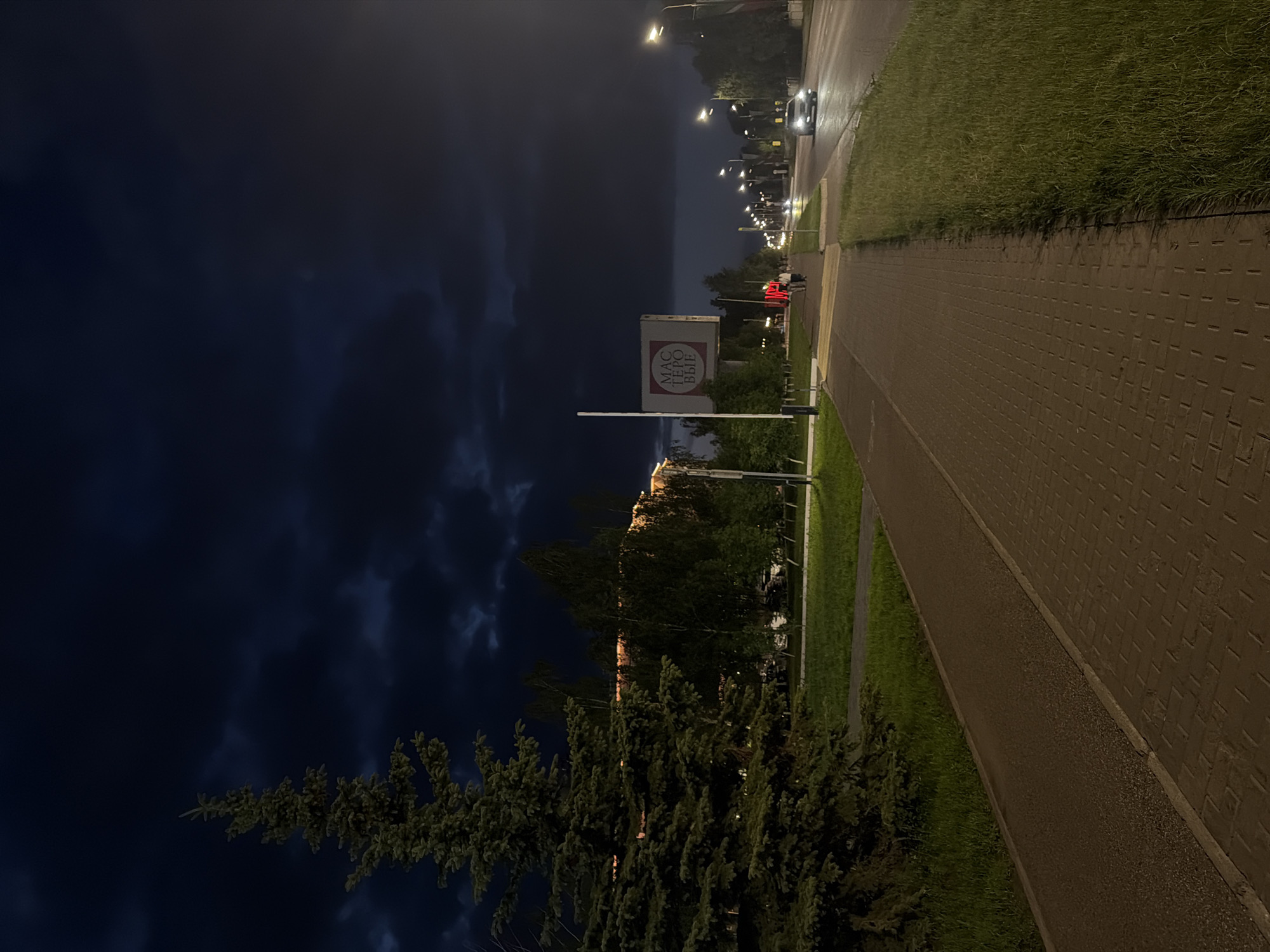 The image depicts a nighttime scene on a city street in Naberezhnye Chelny, Russia, likely during summer due to the green grass and foliage. On the left side of the frame, a large coniferous tree stands prominently, its branches illuminated by distant lights. Adjacent to the tree is a paved sidewalk, followed by a strip of grass and then a road.
The road has a single lane of traffic moving away from the viewer, illuminated by streetlights. A silver car is visible in the right lane, its headlights on. Several streetlights are spaced along the right side of the road, casting pools of light.
On the left side of the road, across the sidewalk and grass, there is a building with lights on its upper floors, suggesting it's a commercial or residential structure. In the middle ground, a large rectangular billboard with red and white lettering is mounted on a pole. The lettering is in Cyrillic and appears to read "МАСТЕРОВЫЕ". Two figures, likely pedestrians, are walking on the sidewalk in the distance, and a red light is visible on a traffic signal or building.
The sky is dark and overcast, with visible dark clouds suggesting a stormy or late evening atmosphere. The overall impression is of a quiet, urban night with signs of ongoing activity.