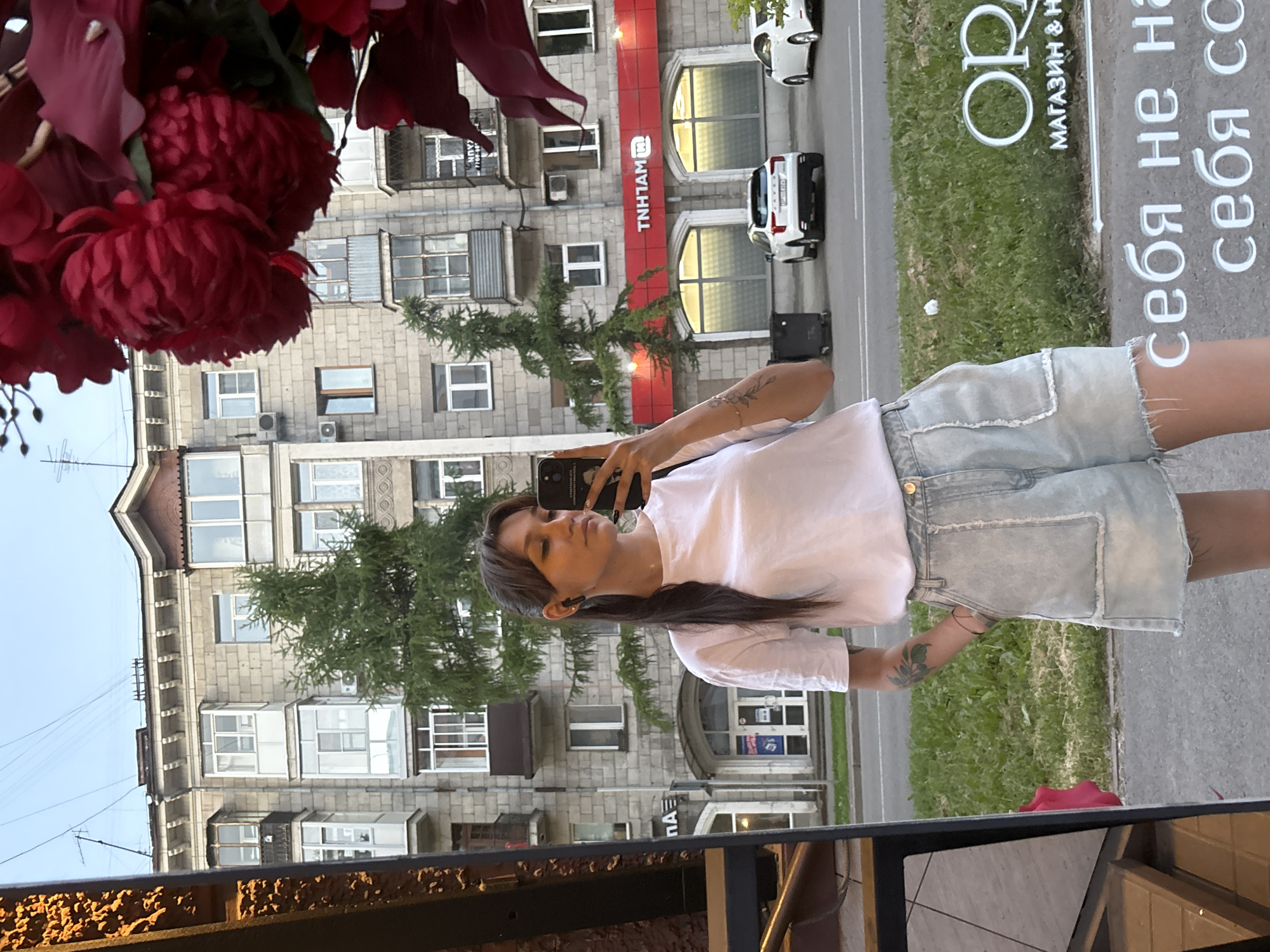 The image shows a young woman standing on a sidewalk, taking a selfie in a reflective surface. She is wearing a white t-shirt and light-wash denim shorts. She has dark hair pulled back into a ponytail and is wearing black wireless earbuds. Behind her, there is a street with cars parked and driving, and a building with multiple windows. A large bouquet of red flowers is partially visible in the foreground. The building appears to be residential or commercial with a red awning that reads "TNHtam". The street sign on the building to the left is partially visible and appears to have "NA" on it. The text on the reflective surface in the foreground reads "себя не на себя со". This scene is located in Novokuznetsk, Russia.