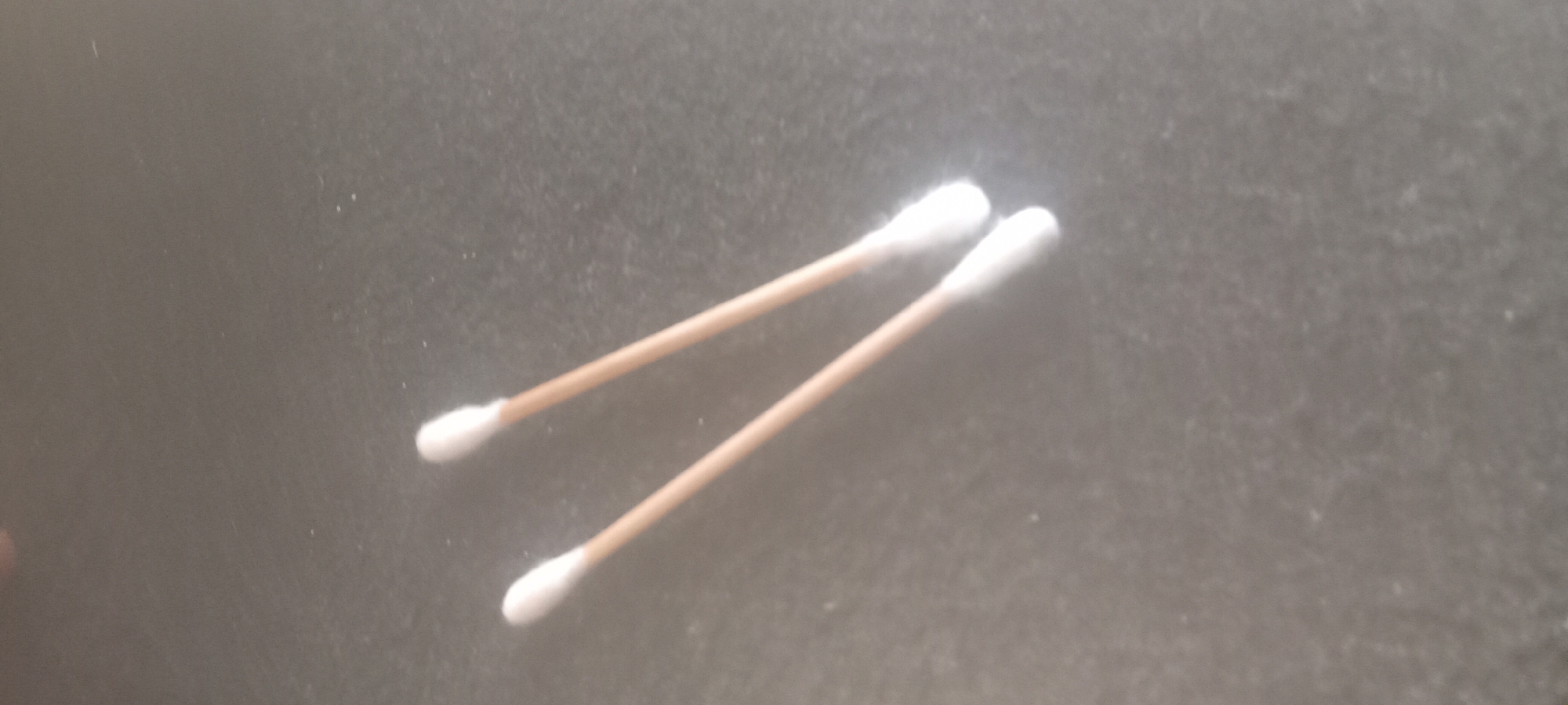Two cotton swabs lie on a dark gray, textured surface. The swabs have light brown sticks and white cotton tips. The surface appears to be slightly dusty or speckled. The location is unknown and there is no indication this picture was taken in Mbribit, Nigeria, or if it contains any people, subjects or actions.