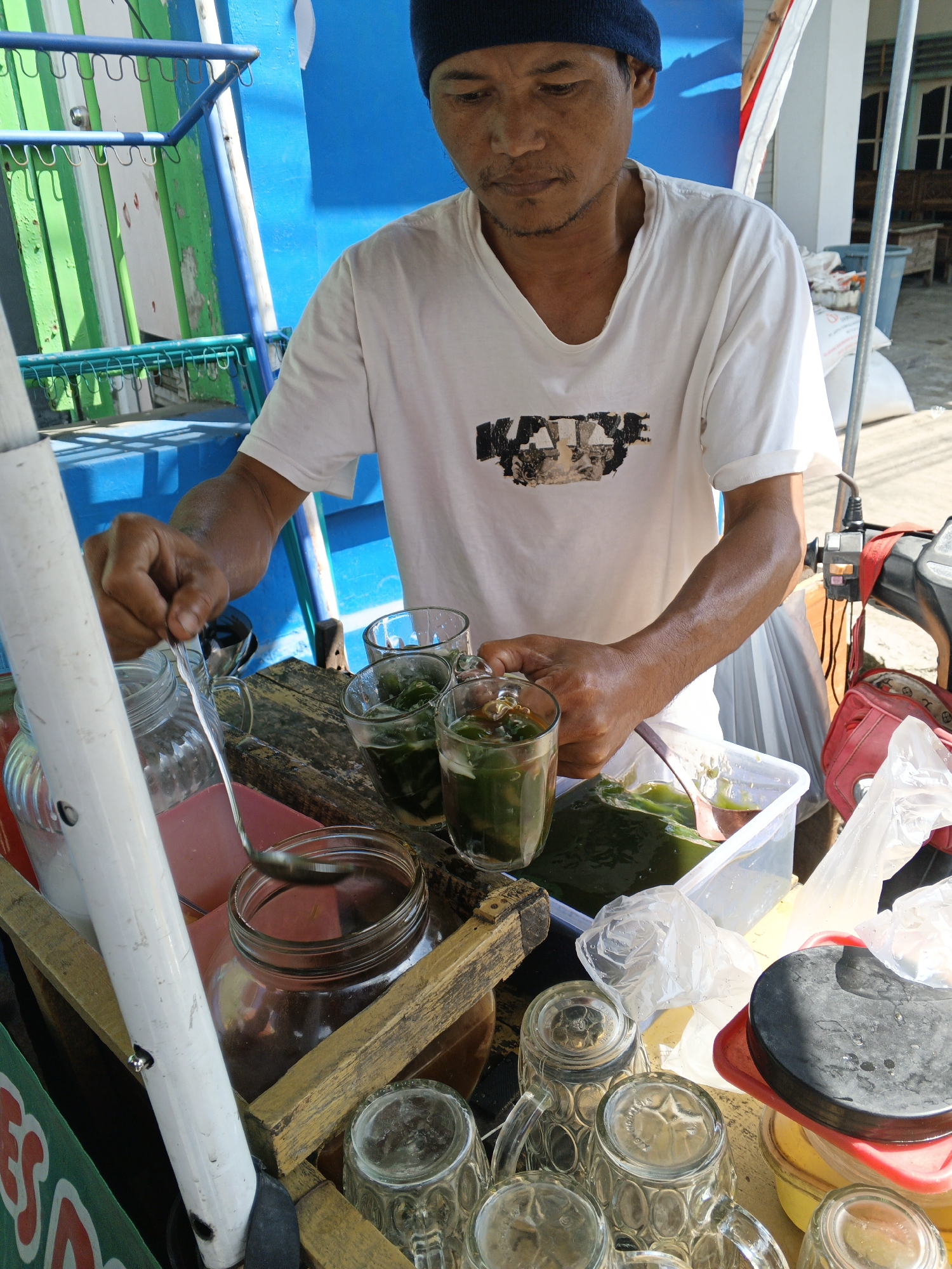 A street vendor in Padang, Indonesia, is preparing drinks. The vendor, a middle-aged man wearing a blue skullcap and a white t-shirt with a graphic print, is seen scooping a green liquid from a plastic container into a glass. The setup appears to be a mobile cart, with various containers, glasses, and a ladle visible. The background suggests an outdoor setting, possibly a street or market, with elements of the cart structure and glimpses of buildings. The overall impression is of a typical day of commerce in a local Indonesian city.
