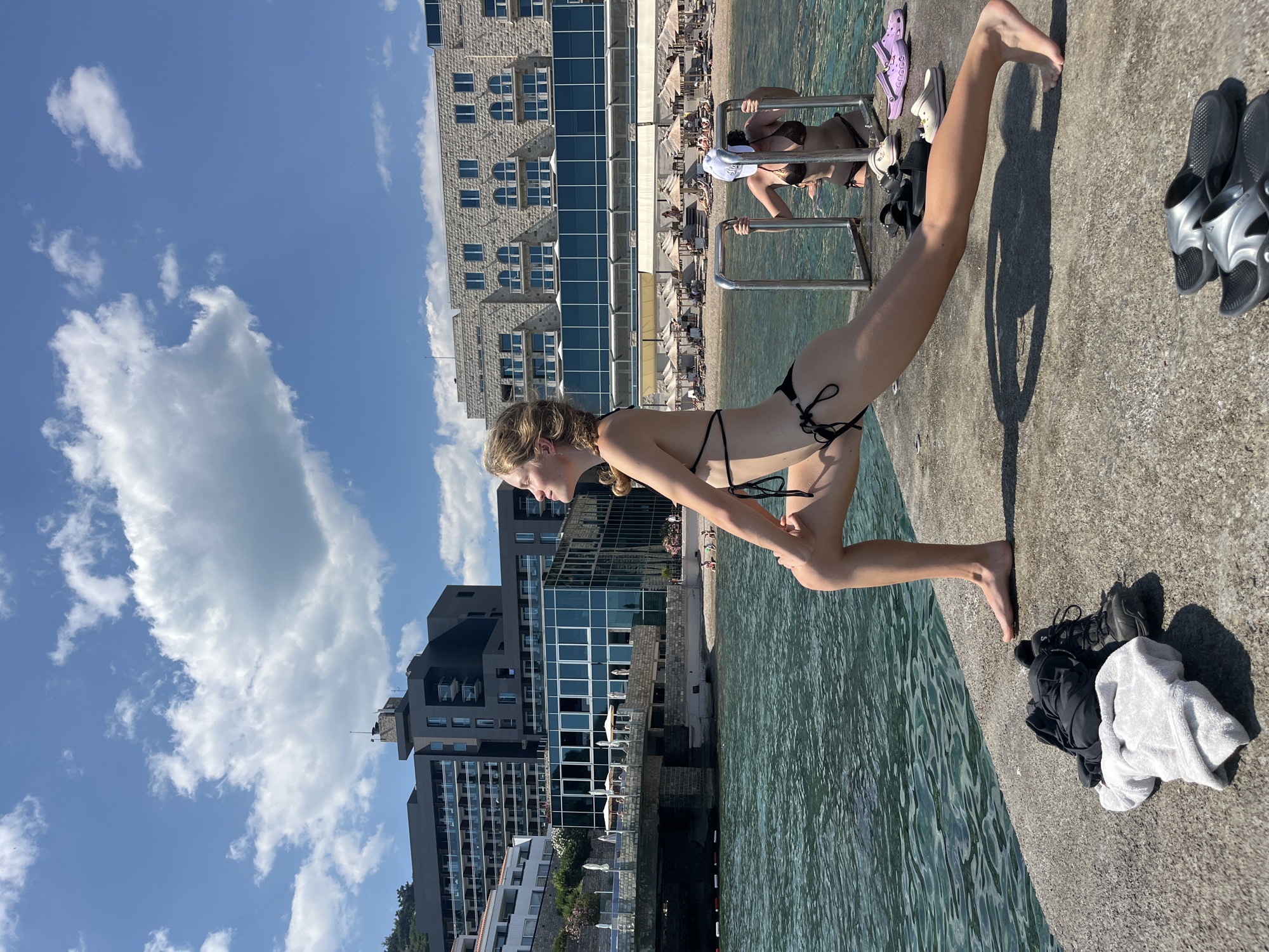 A young woman with blonde hair is doing a lunge exercise on a concrete pier in front of the sea. She is wearing a black bikini. Behind her, another woman in a bikini and a white hat is holding onto a metal railing in the water. In the background, there are large modern buildings and a beach with umbrellas and lounge chairs. The sea is blue and slightly choppy. On the pier, there are a pile of clothes and shoes. The sky is blue with scattered white clouds. The overall scene suggests a sunny day at a coastal resort in Budva, Montenegro.