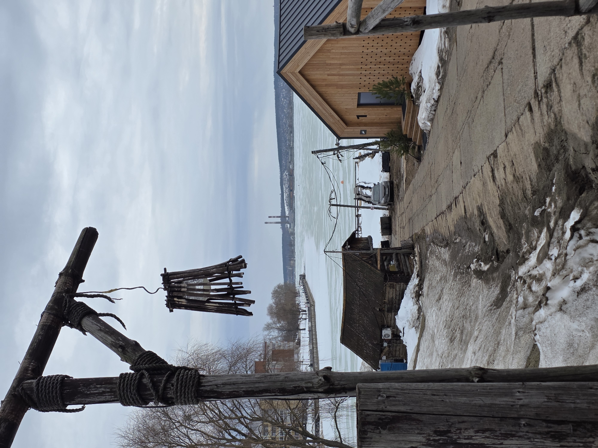 The image depicts a waterfront scene in Votkinsk, Russia, likely in winter. A partially frozen body of water stretches across the middle ground, with industrial smokestacks visible on the opposite shore. The foreground features a wooden structure, possibly a lamp post, constructed from rough-hewn logs tied together with thick rope. A crude wooden lantern hangs from it.
To the right, the edge of a modern, light-colored wooden building is visible. Next to it is a concrete path, partially covered with snow. Further back, a dilapidated wooden structure with a corrugated roof sits closer to the water's edge. A car is parked near this structure. The sky is overcast and the overall atmosphere is cold. The scene contains no people.