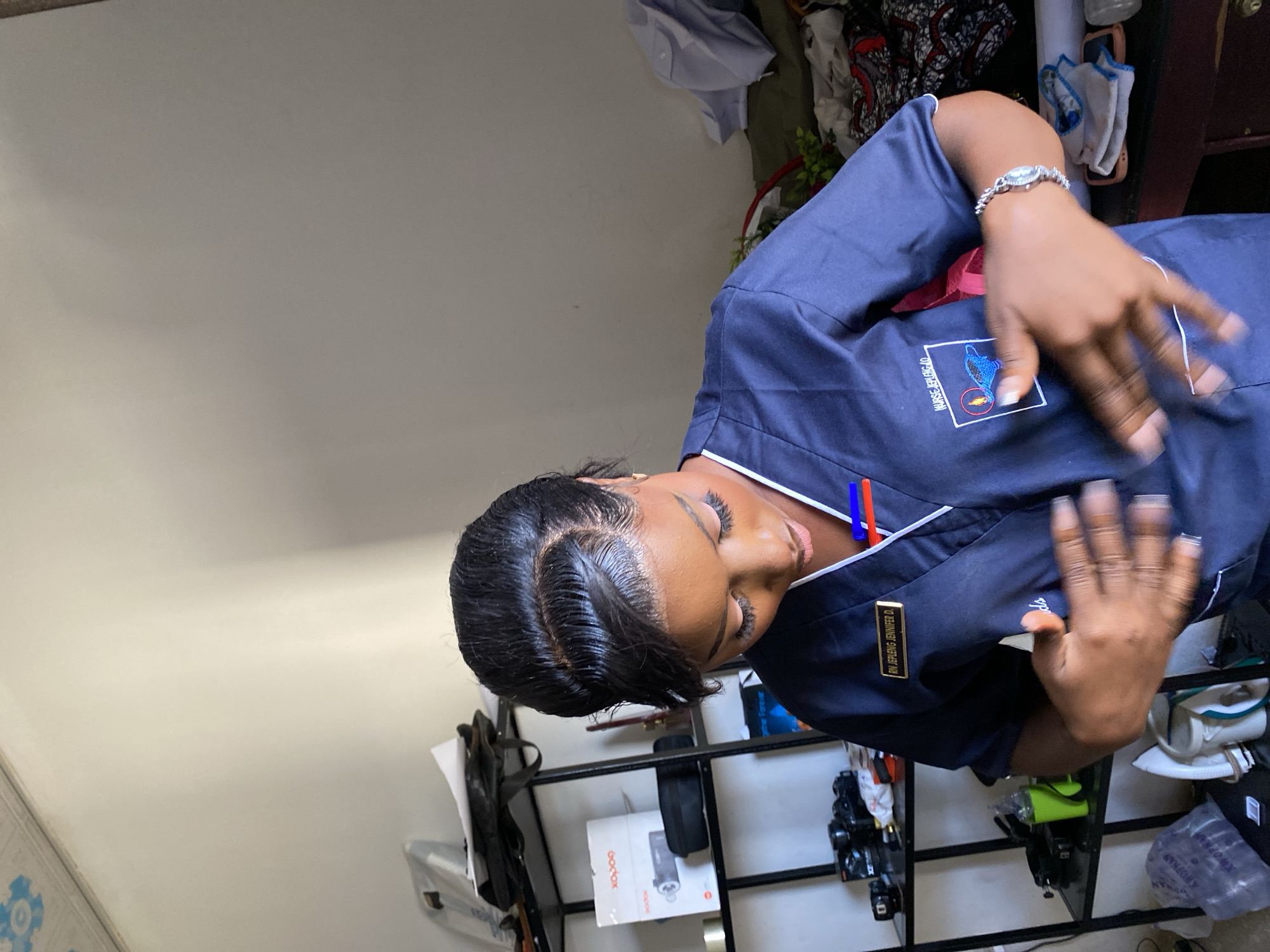 A medium shot of a dark-skinned woman indoors. She has short, black hair with a slight sheen and her gaze is lowered. She is wearing a dark blue nursing uniform with a white trim and an emblem on the chest. A name tag on the uniform reads "RN JENIFER D". She has a watch on her left wrist. Her hands are up with the fingers spread. In the background, there's a white wall and a shelf with photography equipment including a Godox flash, cameras and other miscellaneous items. The location is possibly Gura Top, Nigeria.