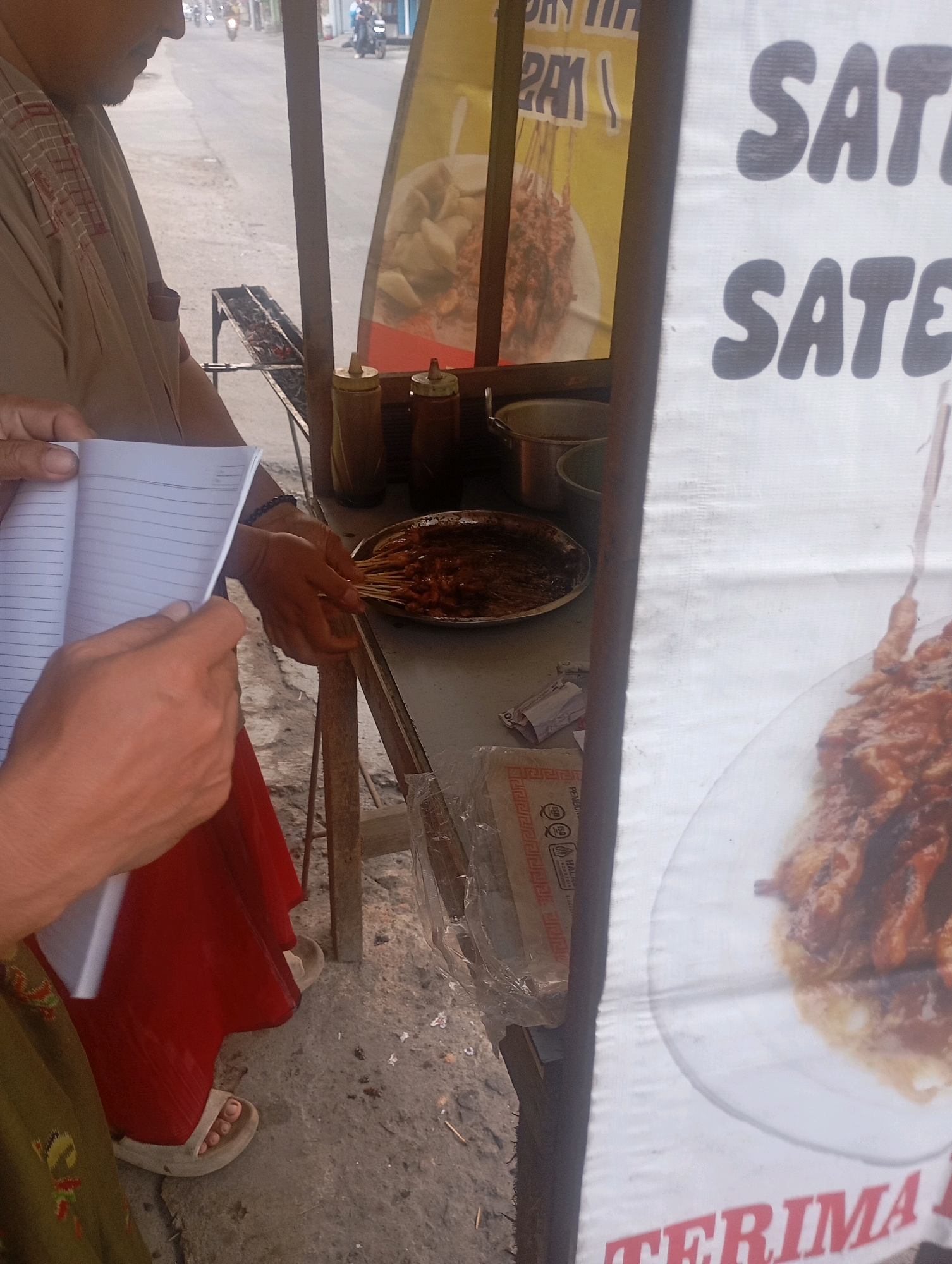 The image depicts an outdoor scene at a food stall, likely in Babelan, Indonesia. A person, partially visible from the side, is wearing a tan shirt with red embroidery and a red skirt. They are holding an open notebook. In front of them, a plate of satay is visible, along with some sauce bottles. A grill with embers is behind the plate. A white banner with large, dark grey lettering reading "SATE" is prominently displayed on the right side of the image. A smaller, yellow banner with text and an image of food is visible behind the grill. The ground is paved with concrete, and there are some visible motorbike tracks in the background, suggesting it is near a road.