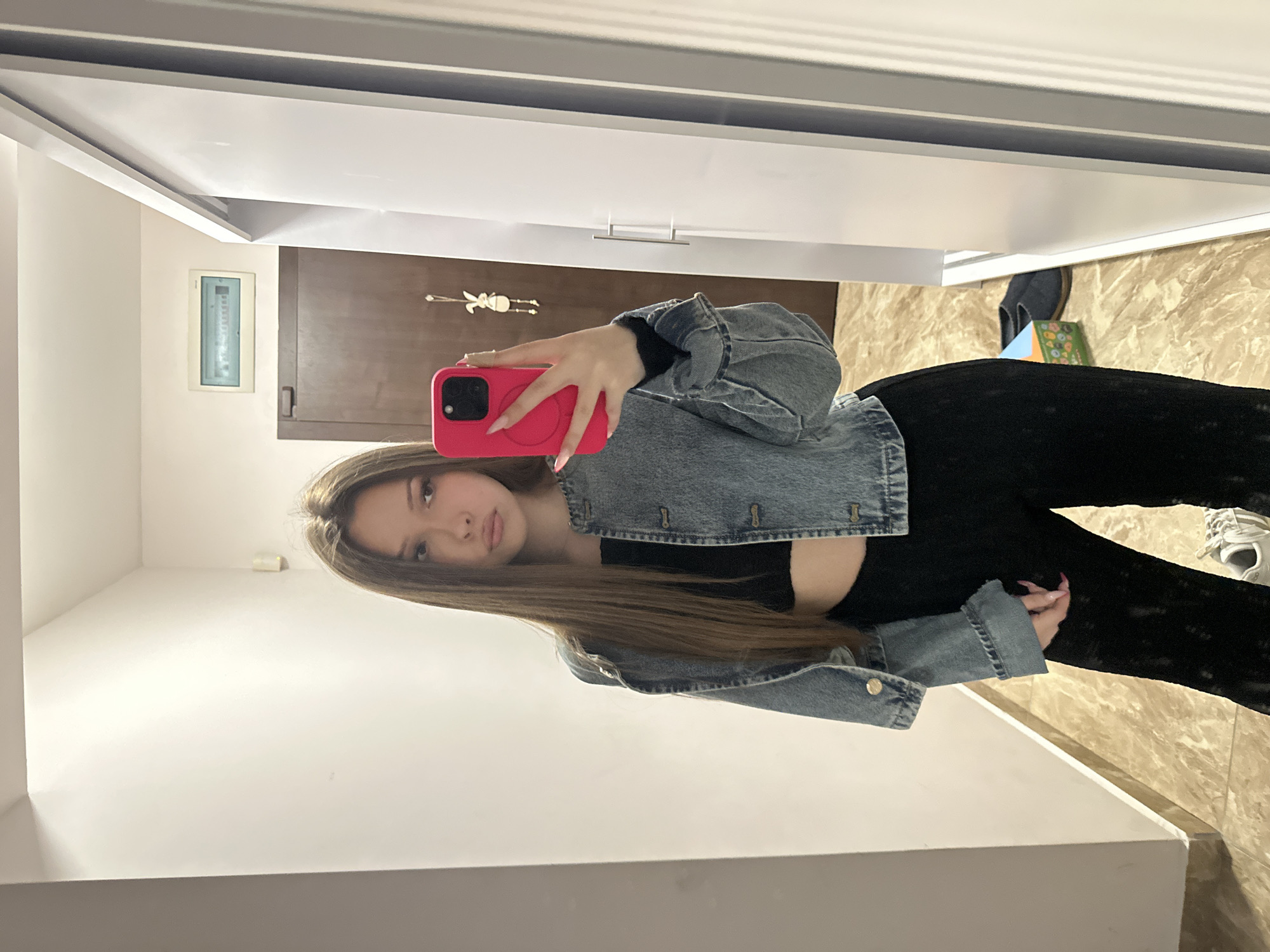 A young woman is taking a selfie in a room. She is wearing a denim jacket, a black top, and black pants. She is holding a pink cell phone in front of her face. The room appears to be a bathroom with a mirror, a door, a wall-mounted electrical panel, and light-colored flooring. The location in the city of Varna, Bulgaria is not discernible from the image.