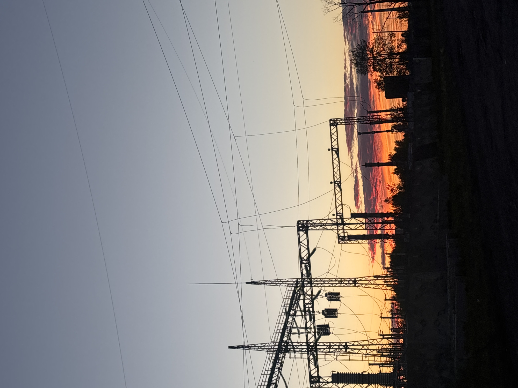 A silhouette of a power substation is visible against a vibrant sunset in Birobidzhan, Russia. The sky displays a gradient of colors, from a pale blue at the top to deep orange and red hues near the horizon, with scattered clouds reflecting the sunset's light. The substation features tall metal towers, complex electrical wiring, and transformers, all rendered in dark silhouette. Trees and other vegetation are also silhouetted along the horizon, interspersed with industrial structures. The foreground is dark, suggesting the image was taken at dusk or dawn, with a rough, unpaved ground surface. There are no people or animals visible in the scene.