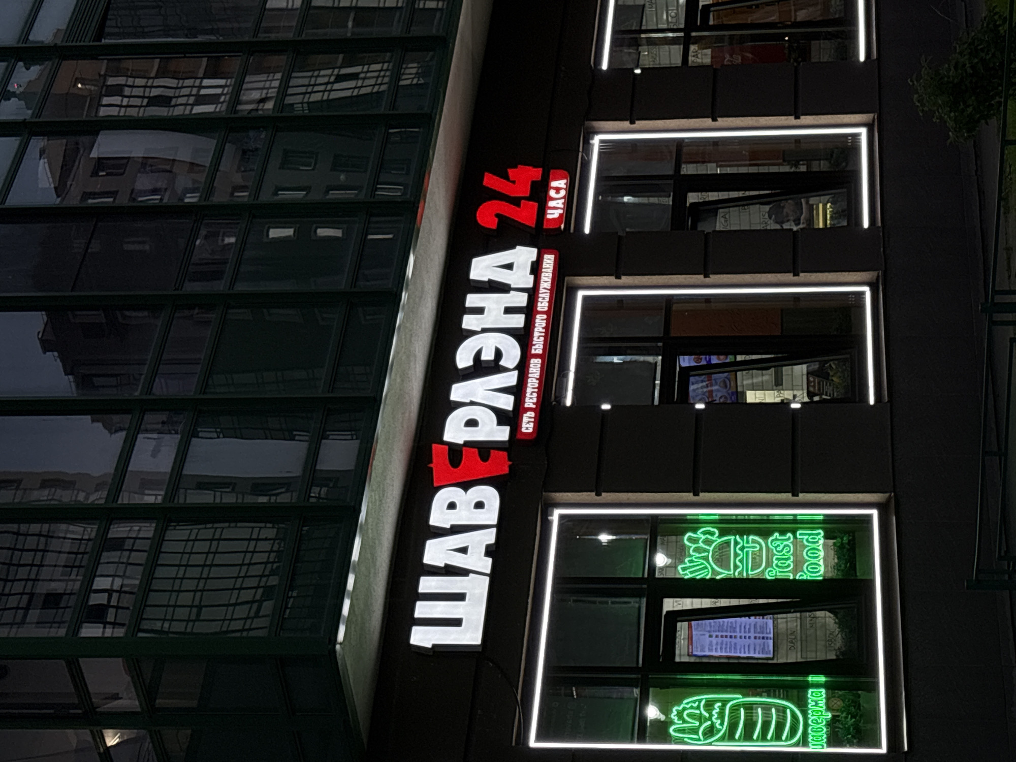 This is a medium-shot, eye-level, nighttime exterior photograph of a brightly lit fast-food restaurant called "ШАВЕРМА 24" in an urban setting. The restaurant has a large, illuminated sign with white block letters that spell out "ШАВЕРМА" with a red stylized "Р" in the middle. To the right of this, the number "24" is in bright red, with the word "ЧАСА" in smaller red letters beneath it. Below the main sign, there is a smaller line of text in white, which reads "СЕТЬ РЕСТОРАНОВ БЫСТРОГО ОБСЛУЖИВАНИЯ".
The storefront features large windows framed by white LED lights. The left window prominently displays a neon green outline of a shawarma and the word "шаверма" in neon green text. Next to it, a menu is visible through the window, with text in Cyrillic. The window to the right of the menu has a neon green outline of a hamburger and the words "fast food" in neon green text. Inside the restaurant, various items are visible through the windows, including what appear to be food displays and shelving. A person is visible in one of the windows in the background, looking down. The building facade above the storefront is dark, with reflections of other buildings in its windows. The ground in front of the restaurant is dark, with some faint lighting. There is some greenery in the lower right portion of the frame. The overall impression is a modern, bustling food establishment operating late into the night.