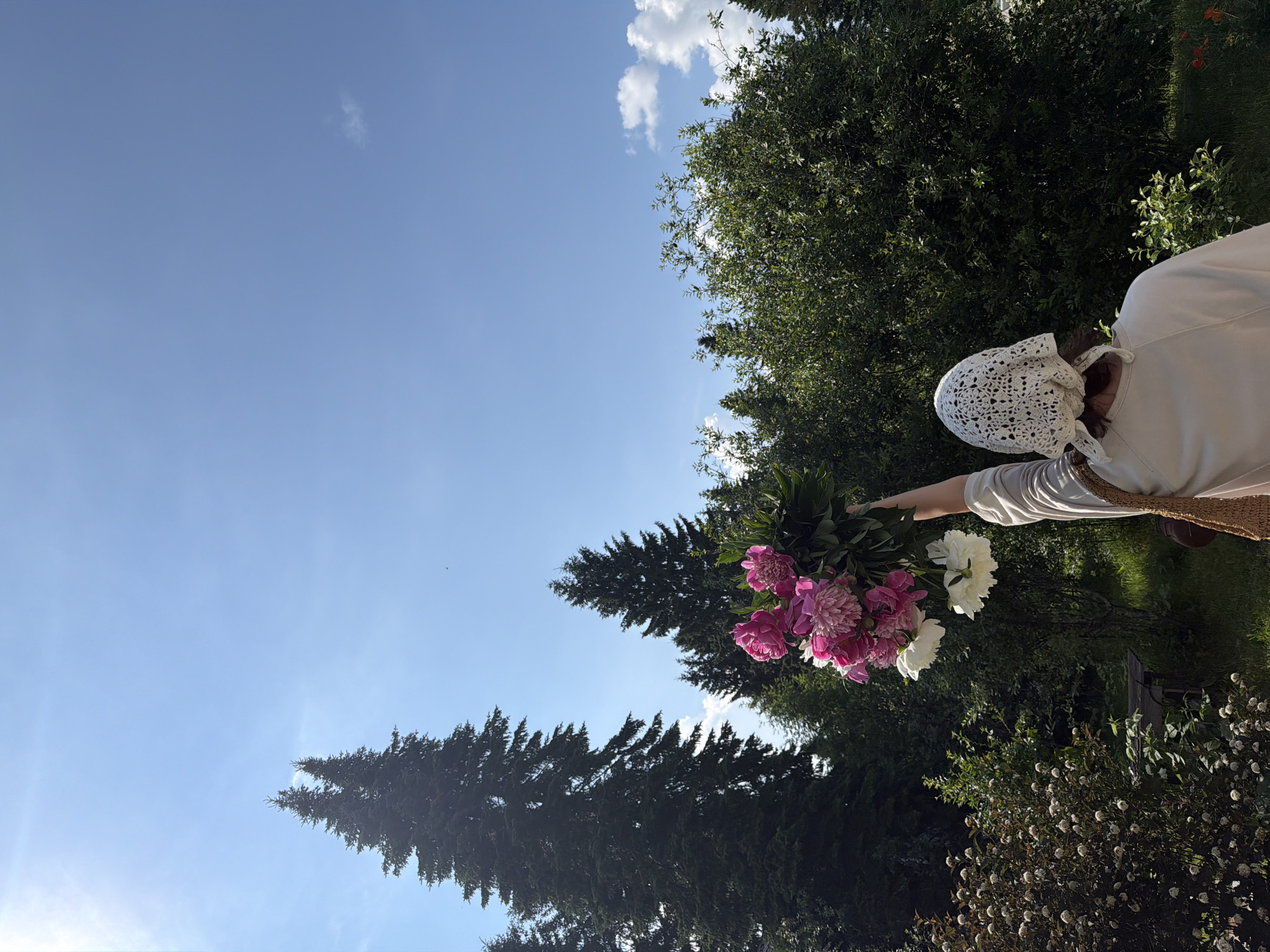 In a natural outdoor setting, a person is holding a bouquet of pink and white peonies aloft. The individual is facing away from the camera, wearing a light-colored long-sleeved shirt and a white crocheted headscarf. Their right arm is extended upwards, holding the flowers. A woven bag is slung over their left shoulder. The background consists of lush green foliage, including tall evergreen trees and other dense vegetation. The sky above is clear and blue with a bright sun, suggesting a sunny day. The location is implied to be the city of Perm, Russia, given the mention of "Прикамский," which is a district within Perm Krai, often associated with the city of Perm and the Kama River region.